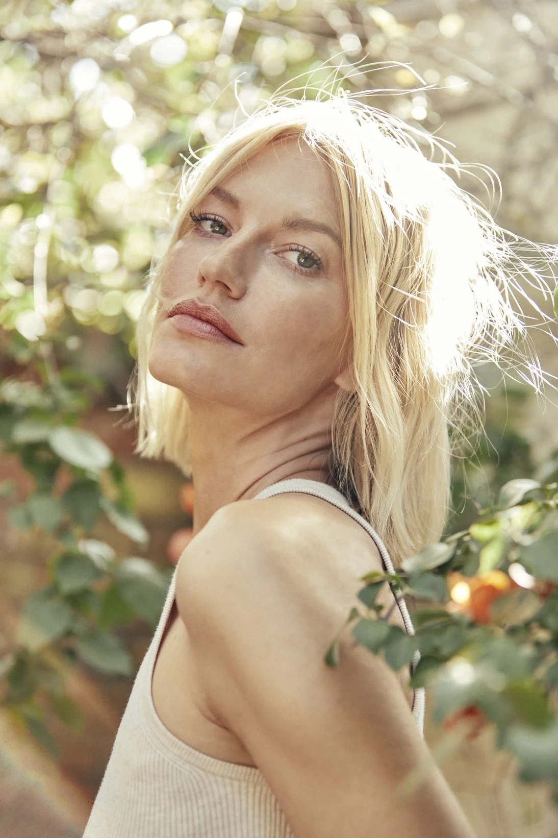 A woman with blonde hair and fair skin outdoors surrounded by green foliage, looking over her shoulder at the camera with a slight smile.