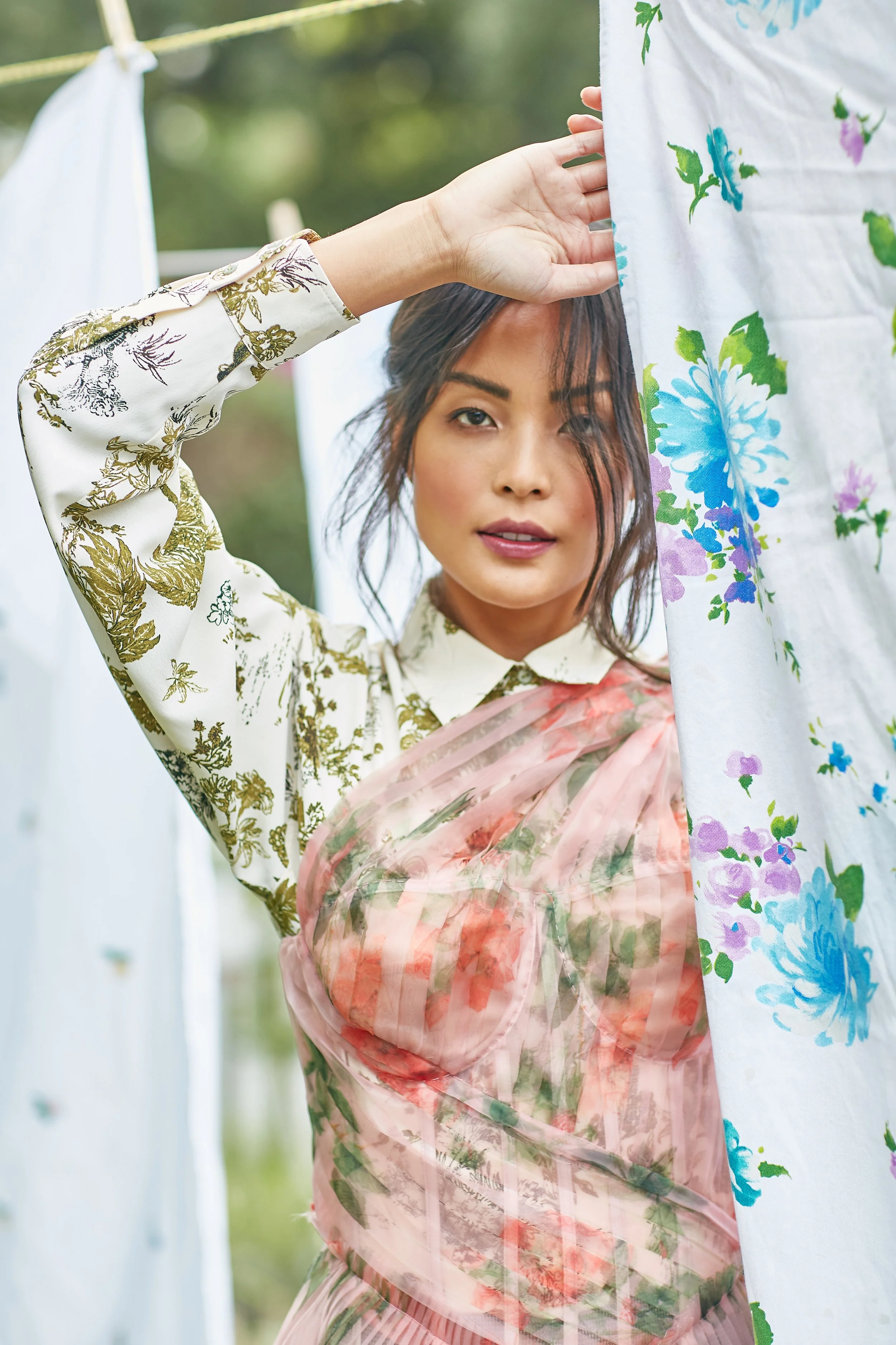 A woman standing outdoors near laundry hanging on a clothesline, wearing a floral top and a sheer floral dress, holding a white floral fabric curtain.
