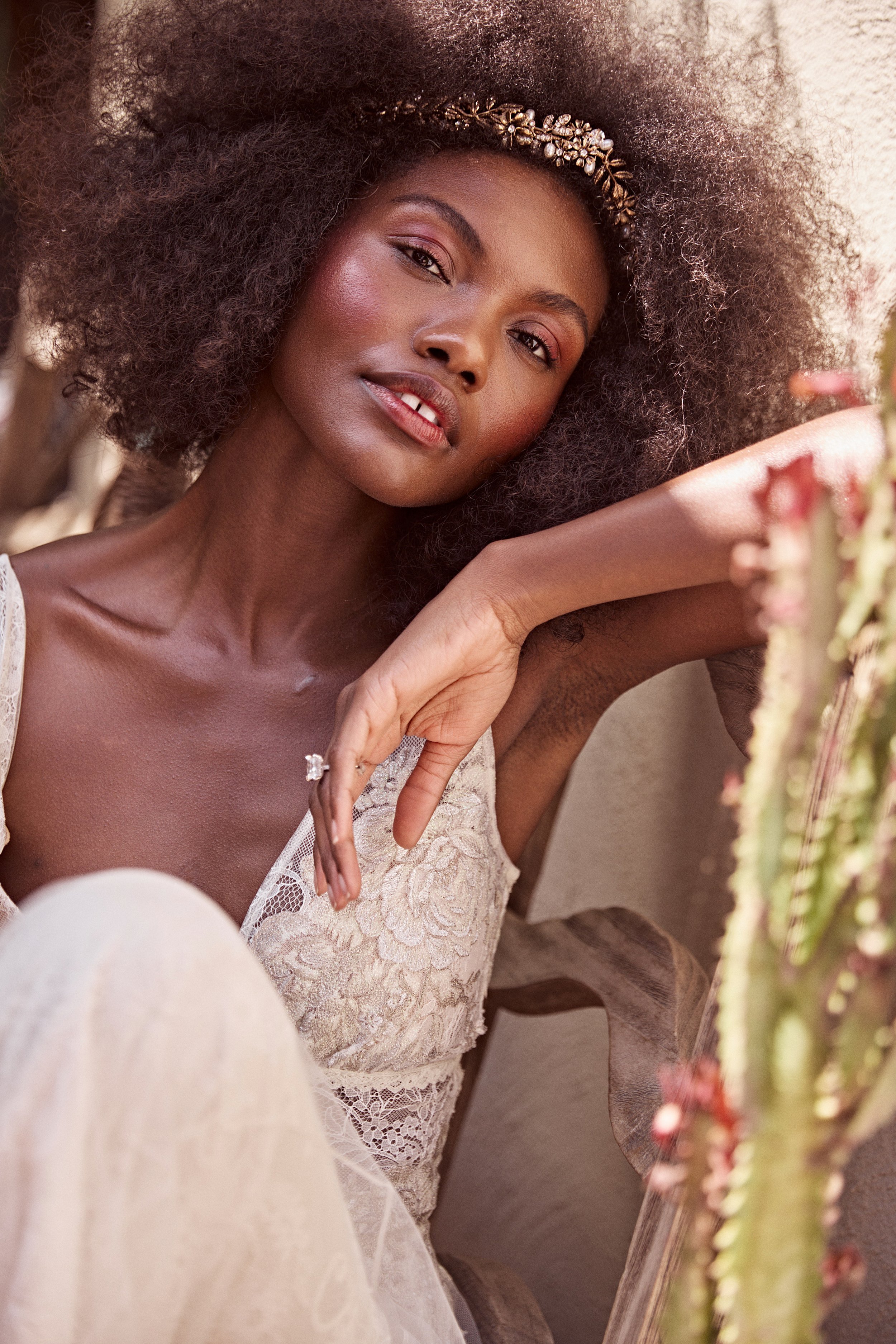 A woman with dark skin and curly hair styled with a decorative headband is sitting on a wooden chair, wearing a beige lace dress. She is resting her head on her arm and looking at the camera with a relaxed expression. The background includes a cactus