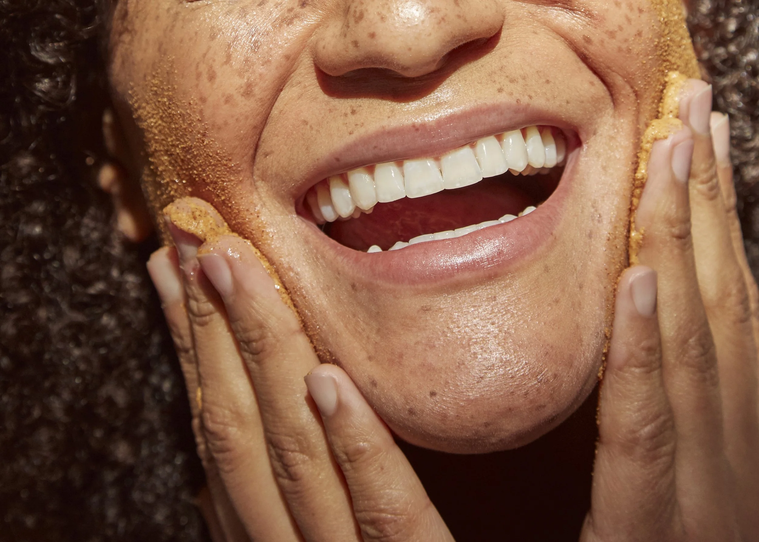 Close-up of a person smiling with white teeth, rubbing face with fingers covered in a brown facial scrub, having curly hair and slightly freckled skin.