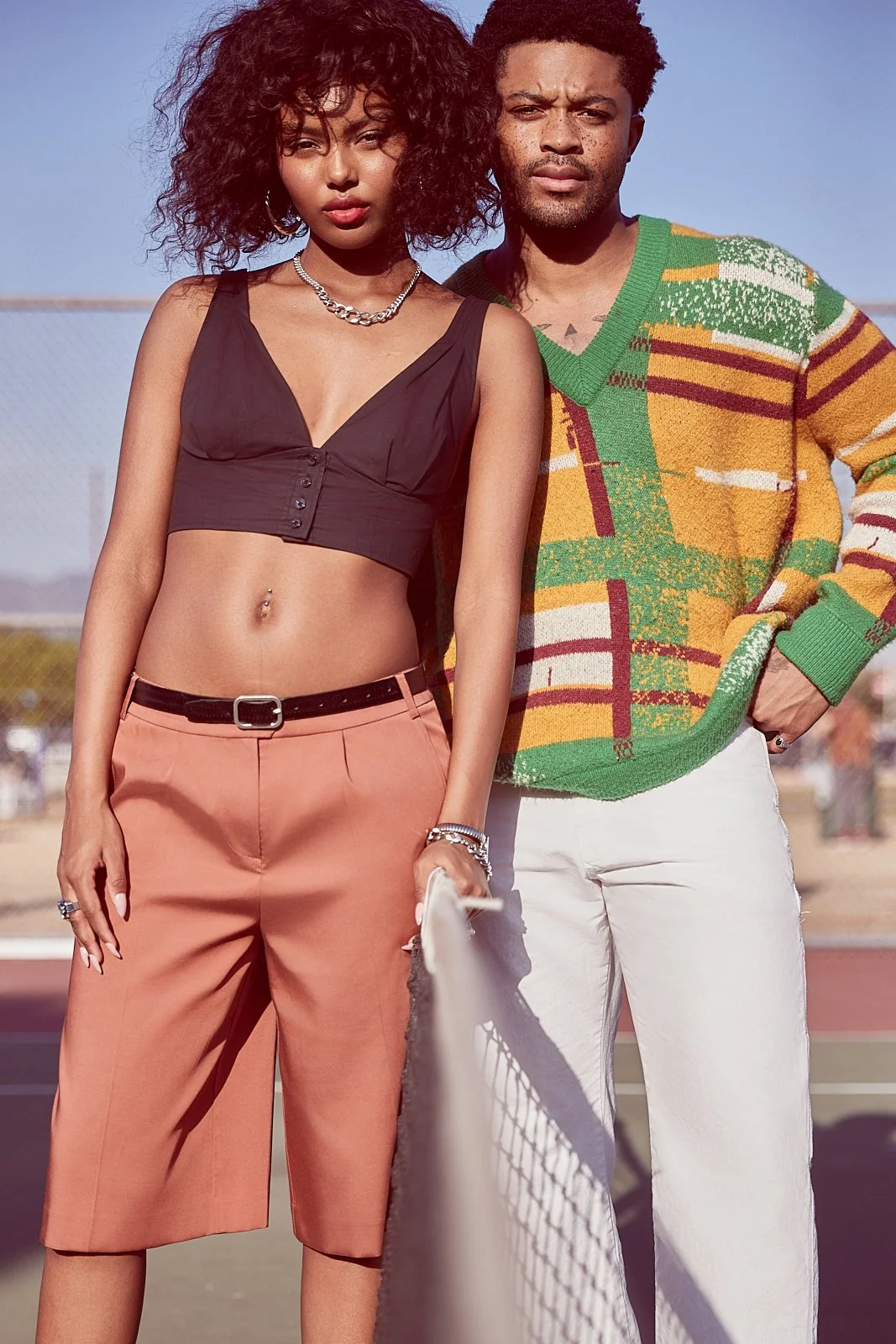 A young woman with curly hair and a man with short curly hair standing on a tennis court, with a clear blue sky in the background; the woman is wearing a black crop top and peach-colored pants, and the man is wearing a colorful sweater and white pant