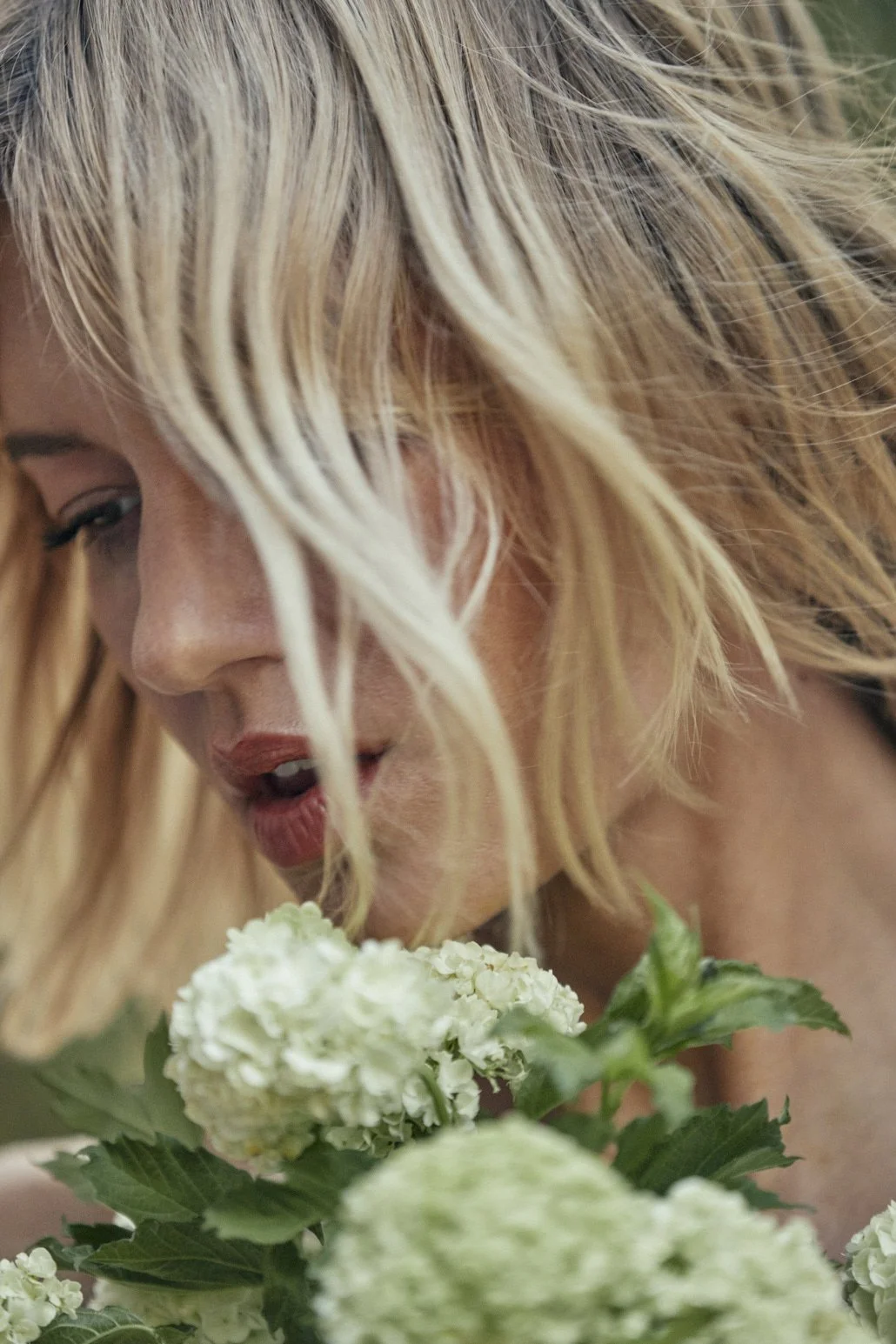 A woman with blonde, tousled hair smelling white hydrangea flowers.