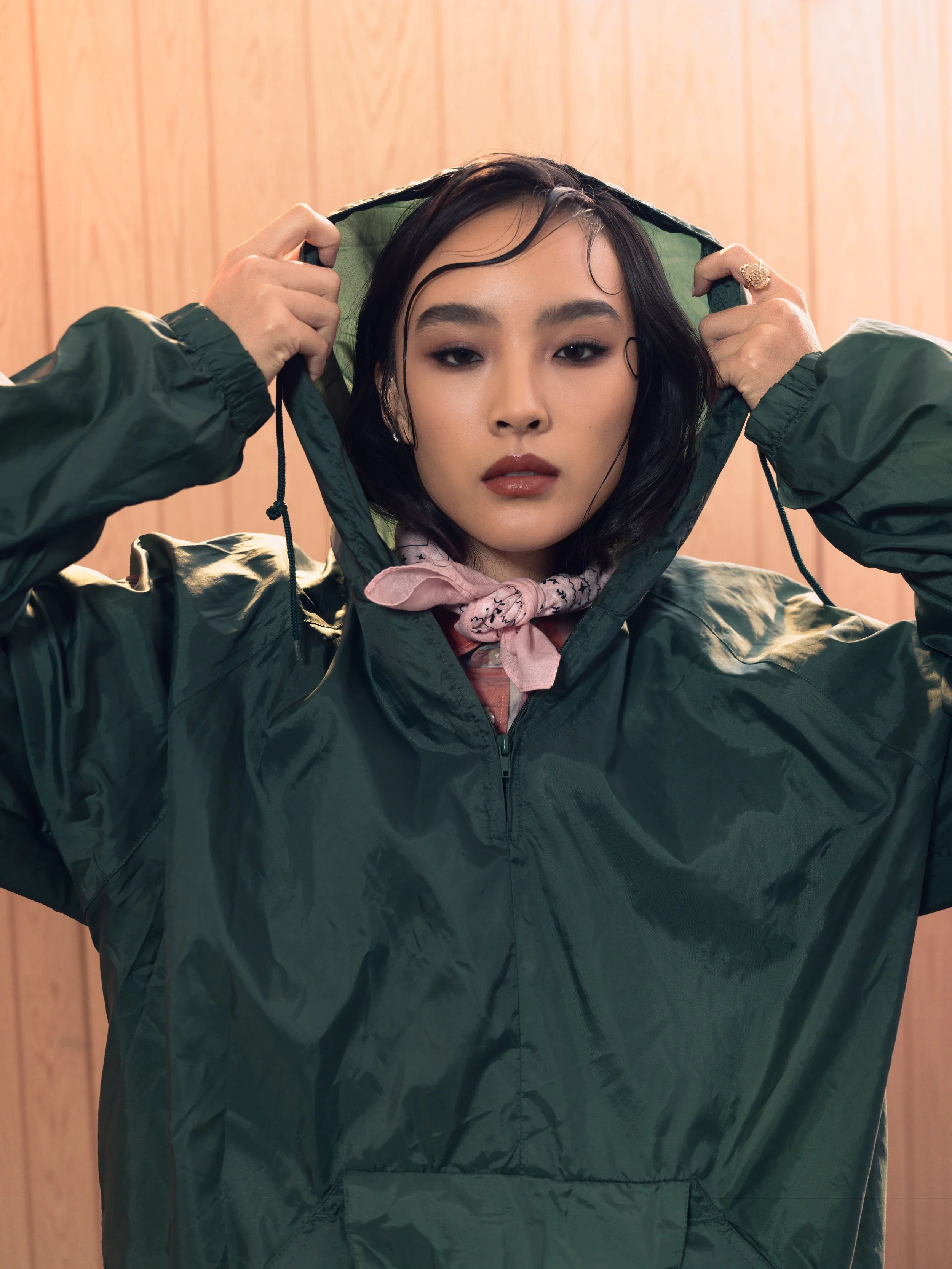 A woman with dark, wet hair wearing a green hooded rain jacket, holding the hood with both hands, standing against a wooden wall.