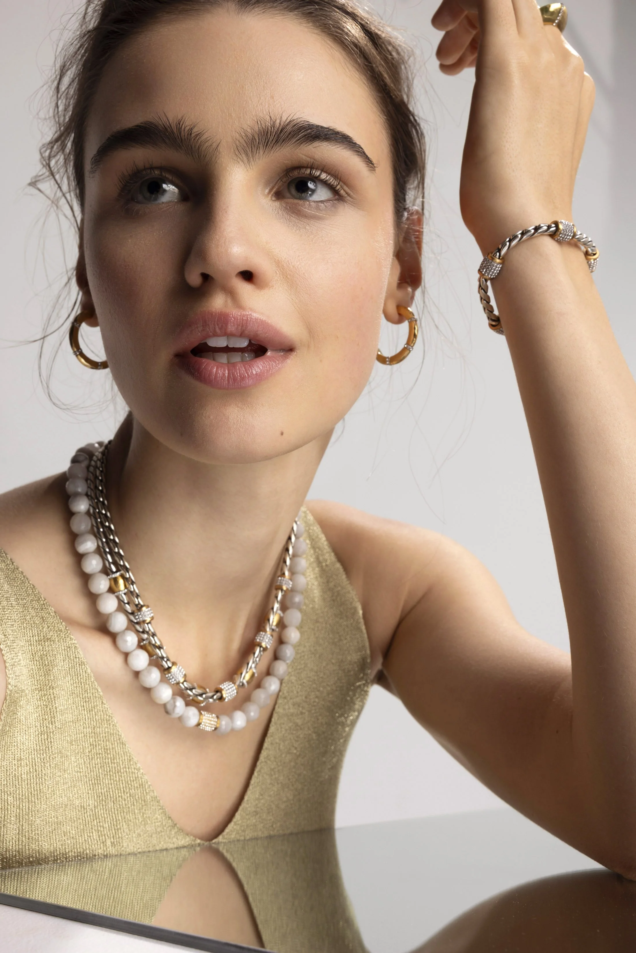 A young woman with light skin, brown hair, and blue eyes wearing gold hoop earrings, layered necklaces with white beads and gold accents, and a gold sleeveless top. She is looking slightly upwards and touching her hair.