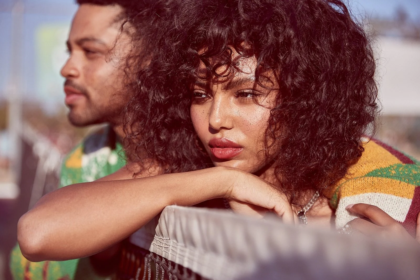 A young woman with curly hair resting her head on her arm, looking contemplative while a man stands beside her, slightly blurred in the background, outdoors with a sunny day.