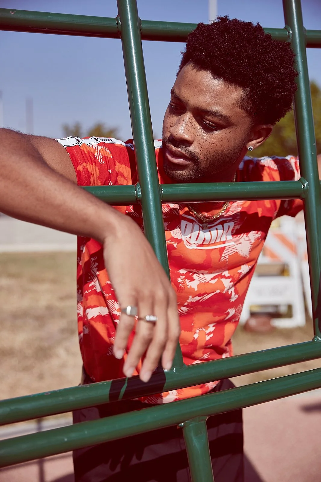 A young man with curly black hair, wearing a red patterned T-shirt, leaning on a green outdoor ladder at a park during daytime.
