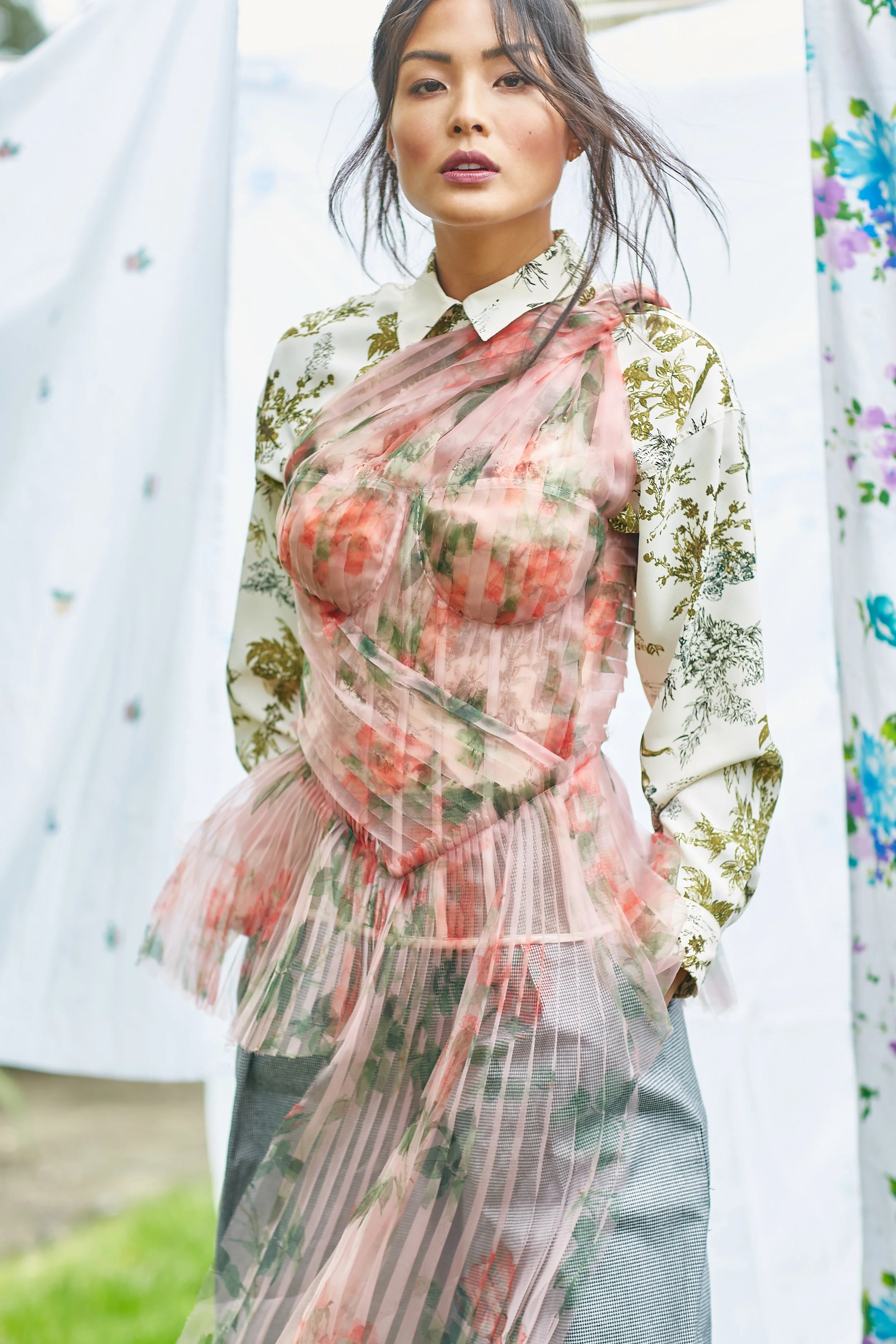 A woman wearing a white blouse with gold floral patterns, layered with a pink and floral sheer dress, standing outdoors in front of white and colorful curtains.
