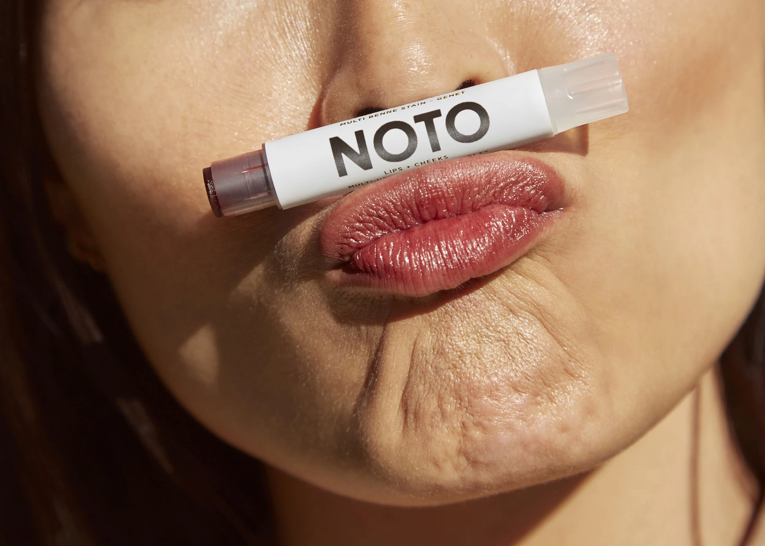 Close-up of a person's lips with a lip balm stick labeled 'NOTO' balanced horizontally on the lips, with visible skin texture and natural lip color.