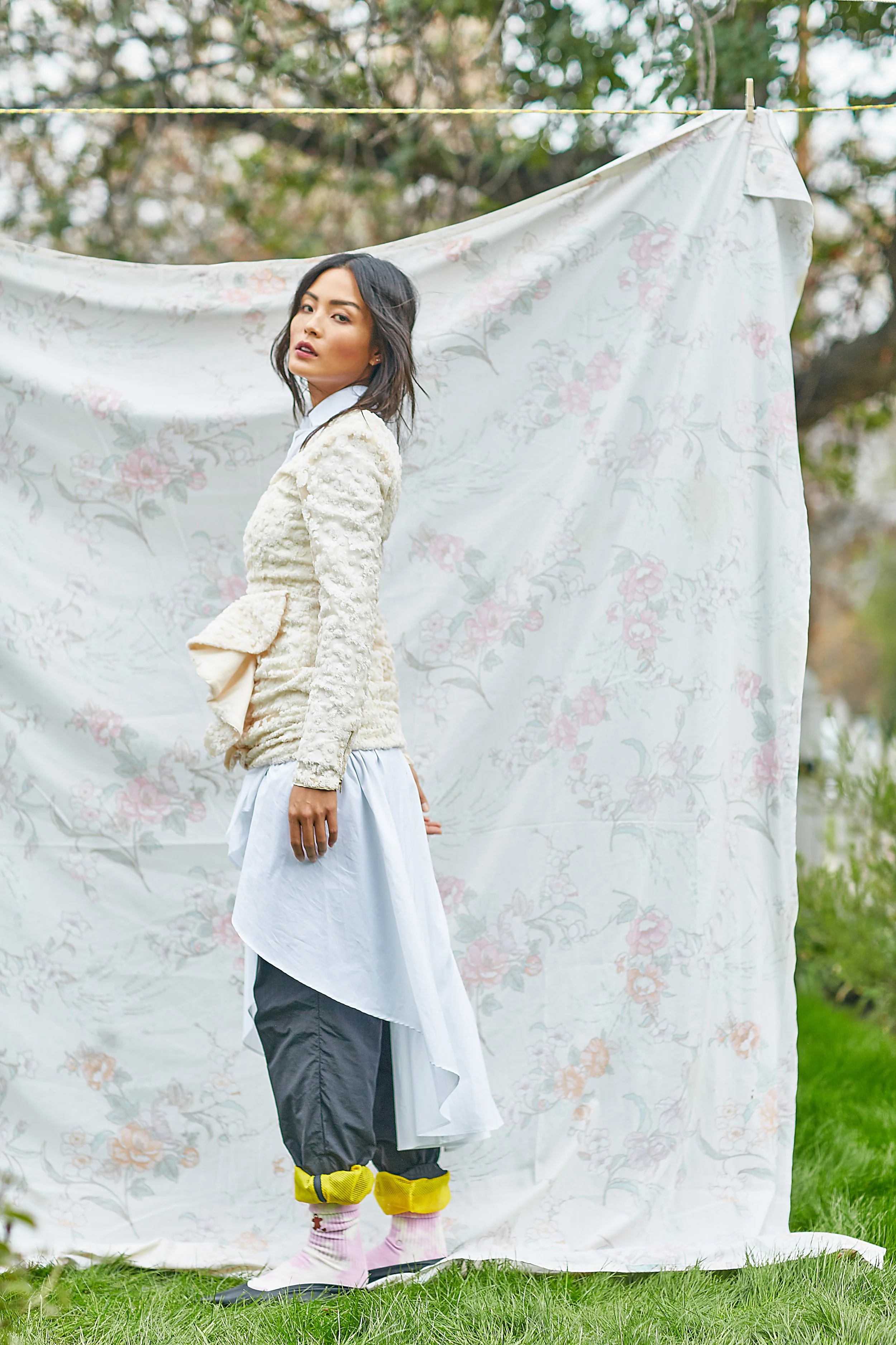 A woman standing outdoors in front of a floral backdrop, wearing a textured cream-colored jacket, layered with clothing, and colorful socks.