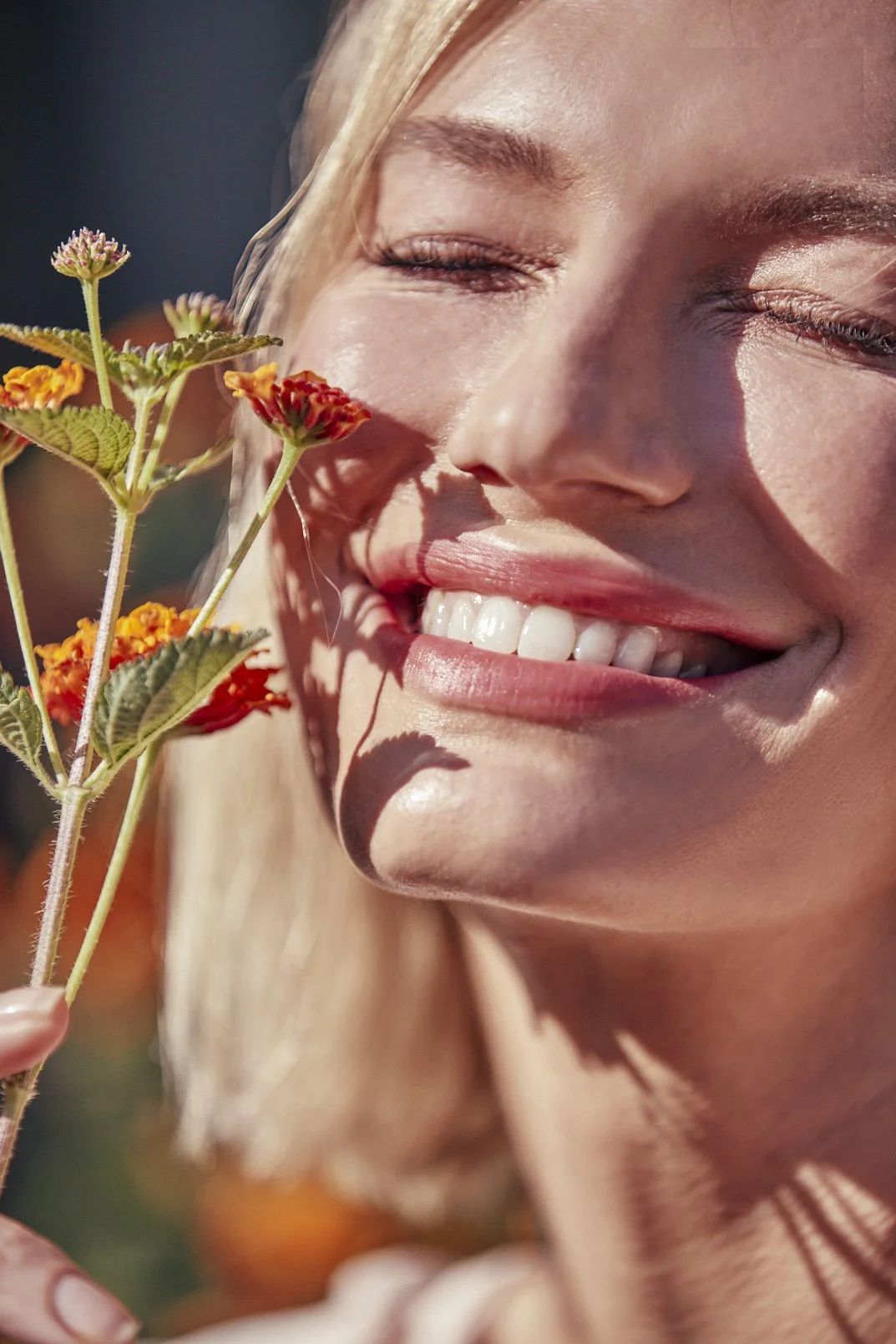 A smiling woman with closed eyes holding a small colorful flower close to her face in sunlight.
