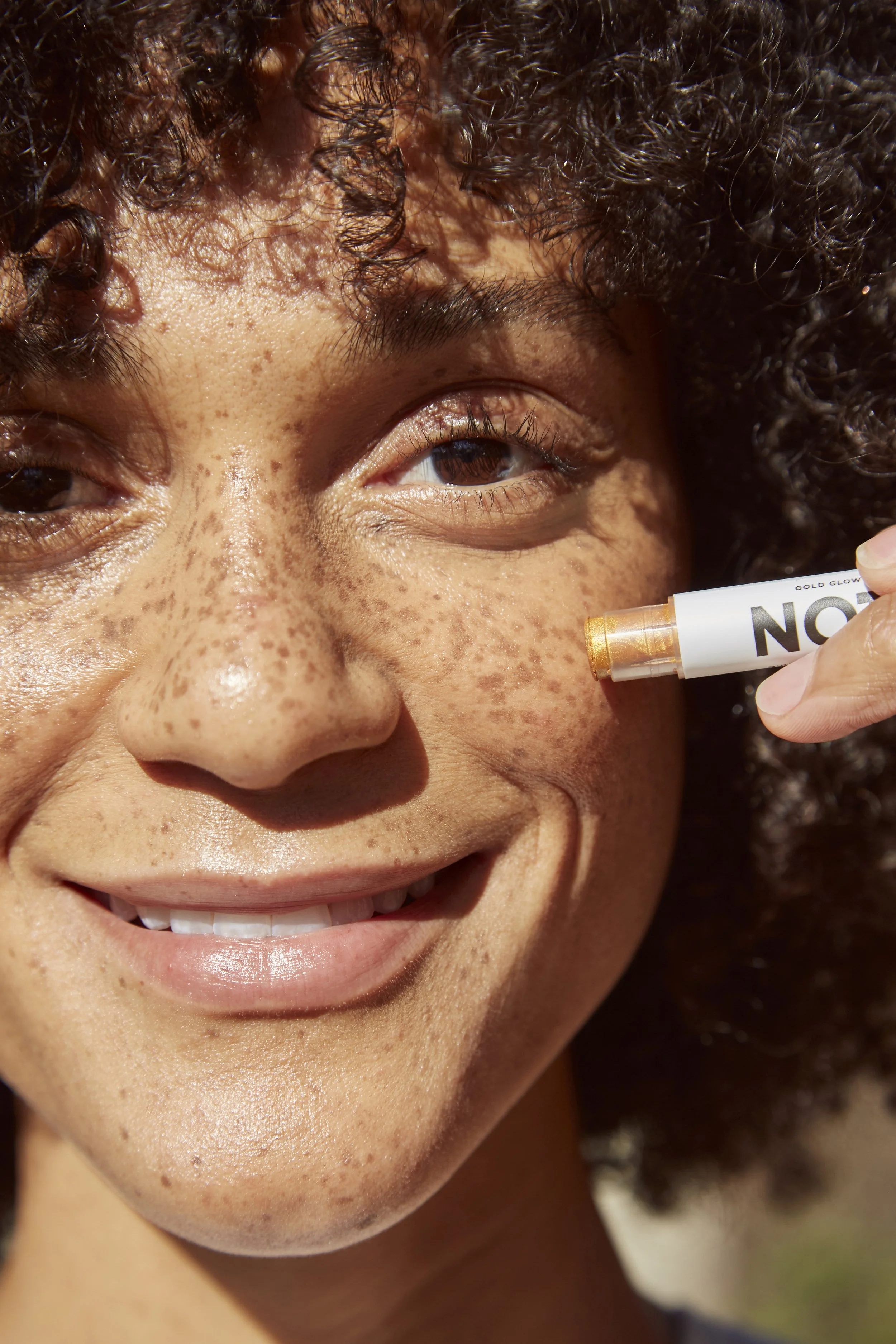 Close-up of a woman with curly hair and freckles smiling while applying a shimmering gold highlighter to her cheek with a stick marker.
