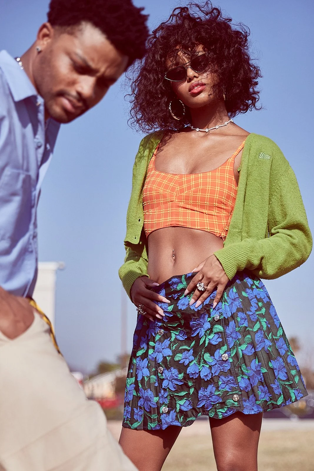 A woman wearing sunglasses, an orange plaid crop top, a green cardigan, and a floral skirt stands outdoors under a clear blue sky, with a man partially visible in the foreground.