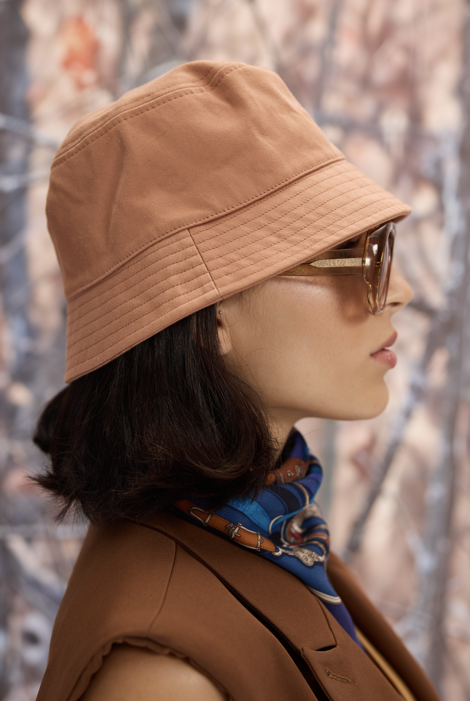 Side profile of a woman wearing a light brown bucket hat, glasses, a blue and brown scarf, and a brown jacket with a background of blurred autumn trees.
