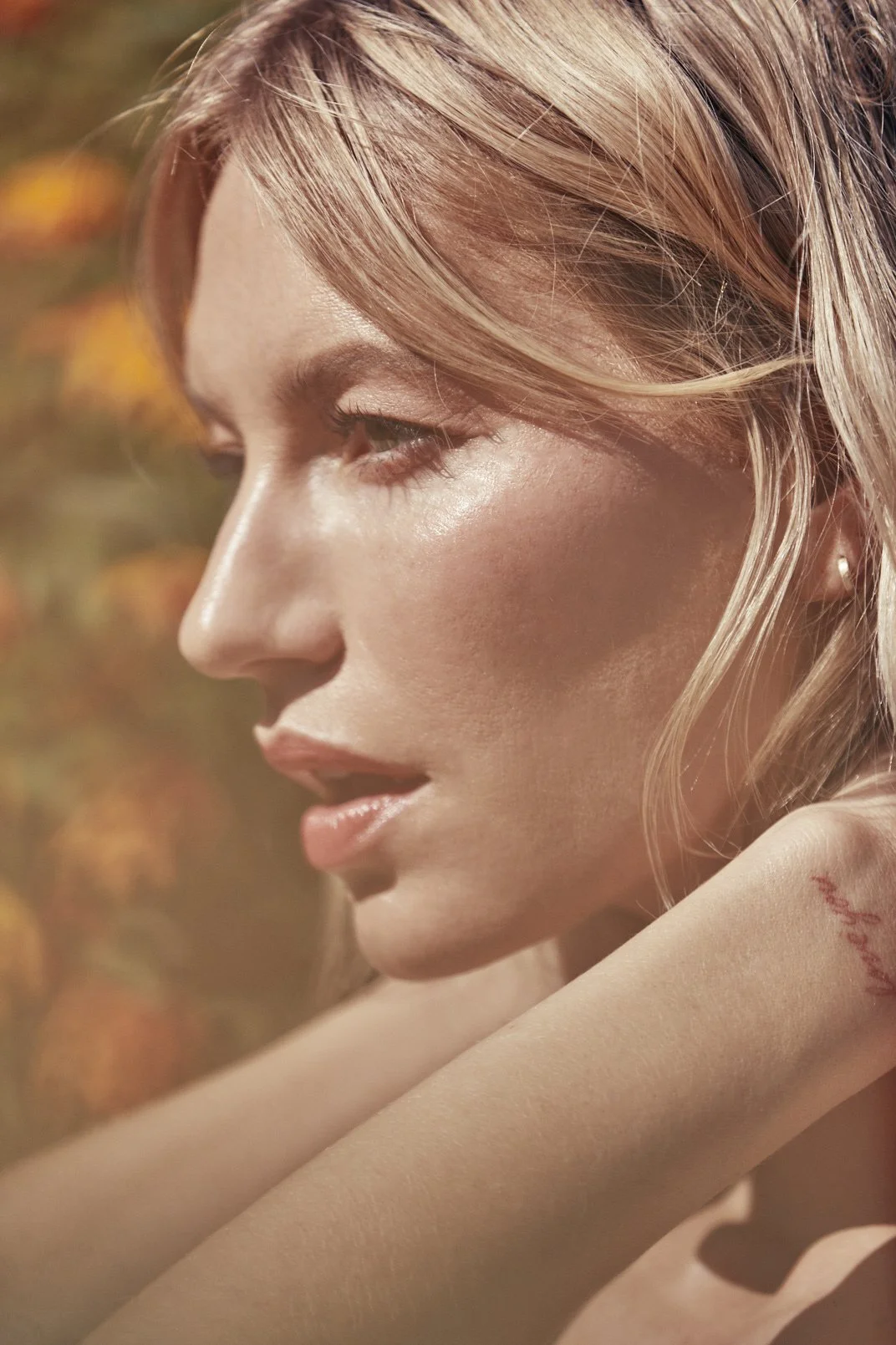 Close-up side profile of a woman with blonde hair, smooth skin, and a visible tattoo on her arm, outdoors with a blurred autumn-colored background.