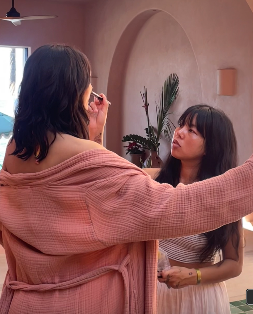 Two women are indoors, one applying makeup or lipstick to the other. The woman on the left has shoulder-length dark hair and is wearing a pink textured bathrobe. The woman on the right has long dark hair with bangs and is wearing a sleeveless striped top and a light-colored skirt, holding a small object in her hand. The background features a pinkish wall with a potted plant and natural light coming through a window.