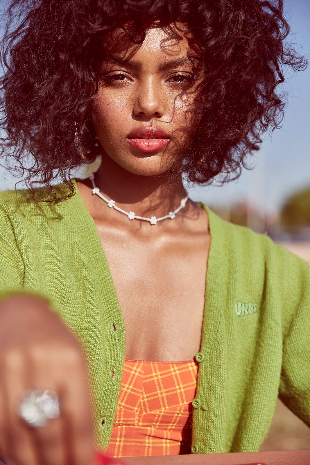 Close-up of a woman with curly hair wearing a green cardigan, orange top, and a white flower necklace, outdoors in daylight.