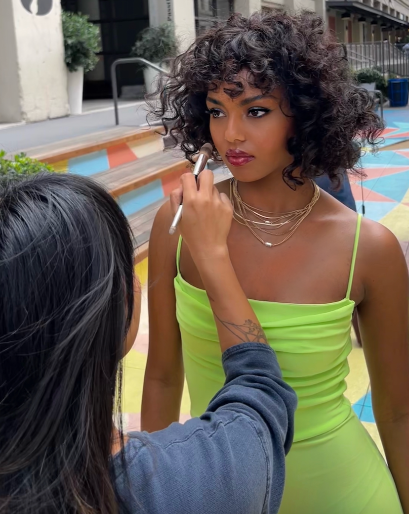 A woman with short curly hair and layered necklaces is getting her makeup done outdoors. The makeup artist, with long dark hair, is applying makeup with a brush. The woman is wearing a neon green dress with thin straps, and the background features steps with colorful paint patterns.