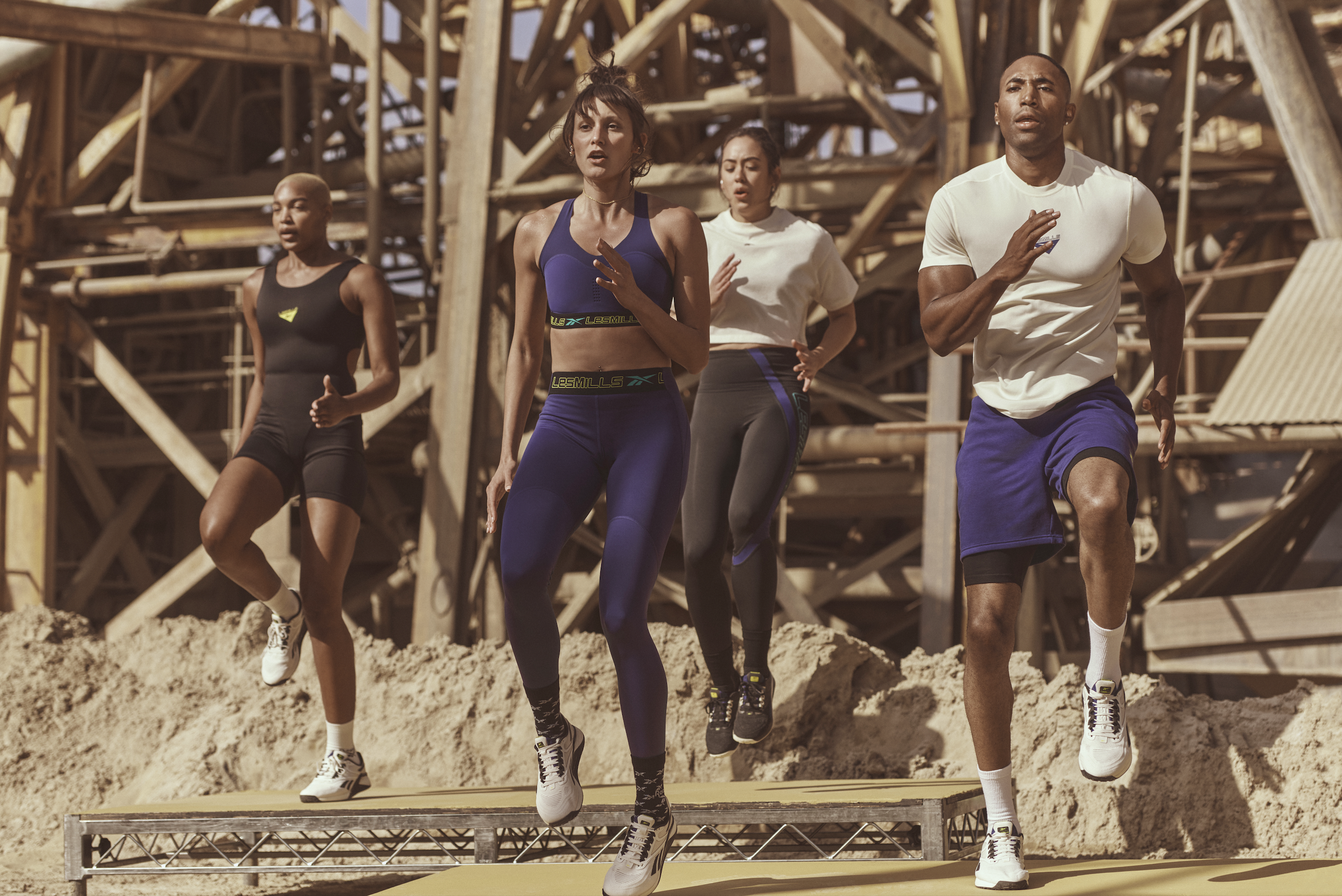 Four people running outdoors on a sandy surface with a wooden structure in the background.