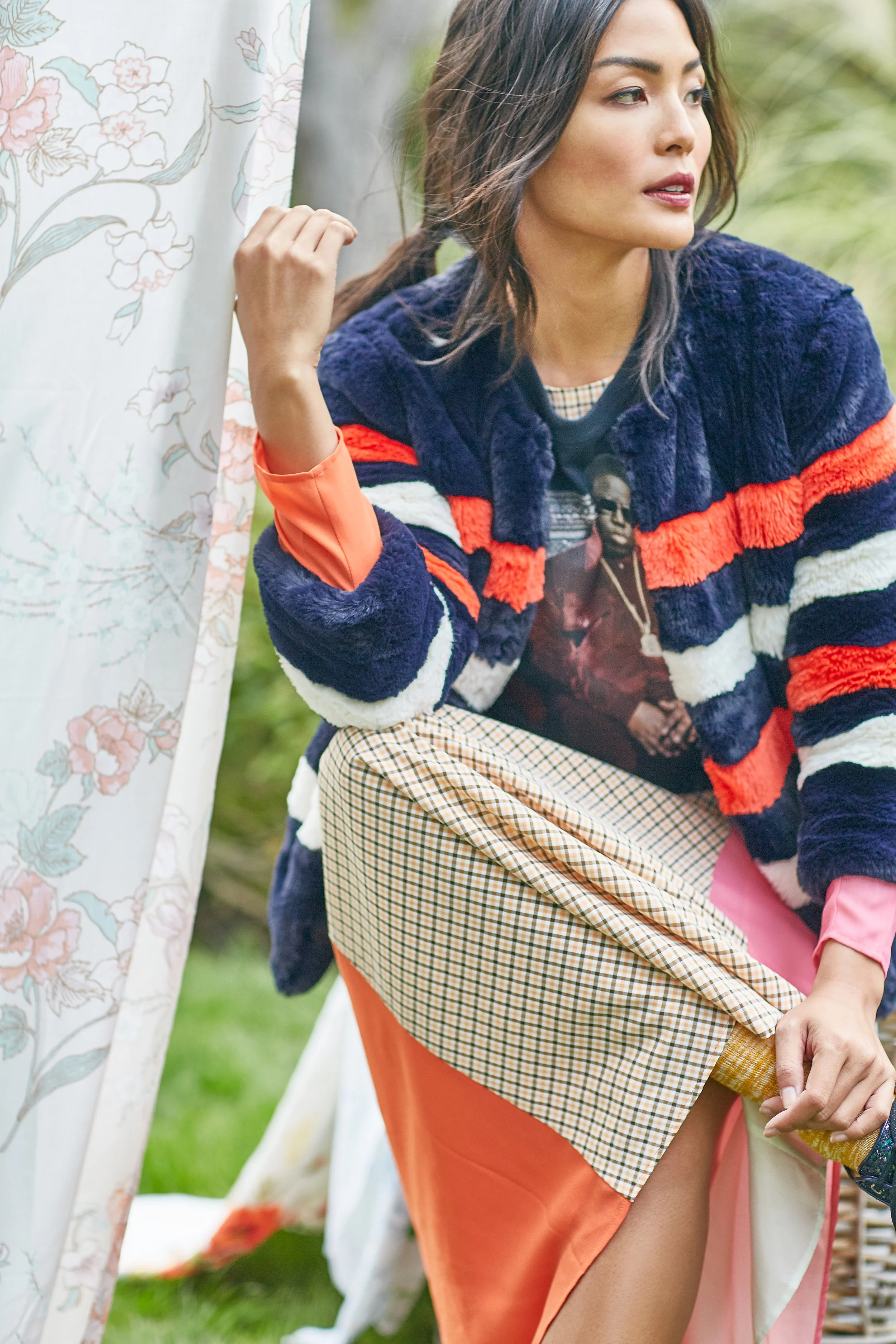 A woman with dark hair and makeup, wearing a multicolored striped jacket over a graphic T-shirt, sitting outdoors near a floral curtain.