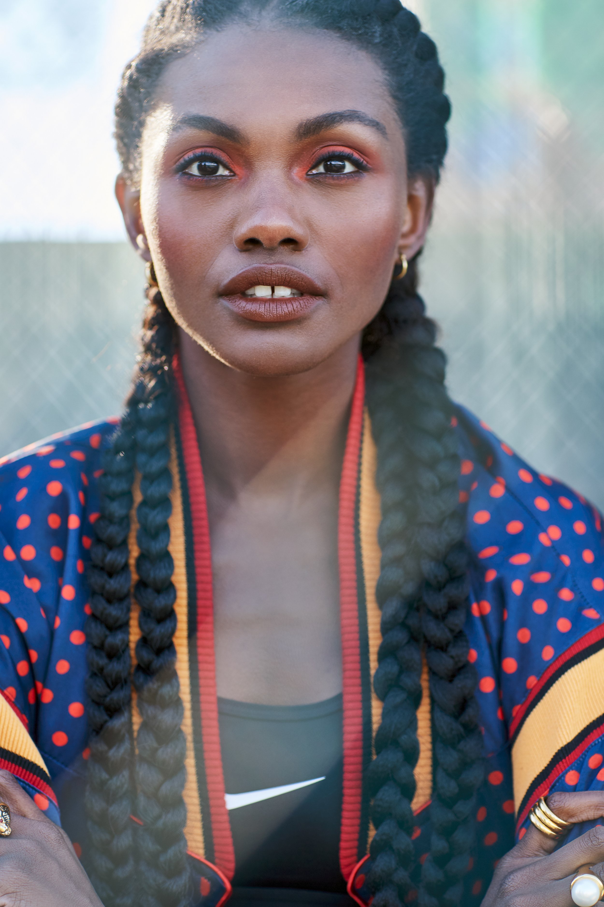 A woman with dark braided hair, wearing a sports top under a colorful, patterned jacket, adorned with jewelry, outdoors in natural light.