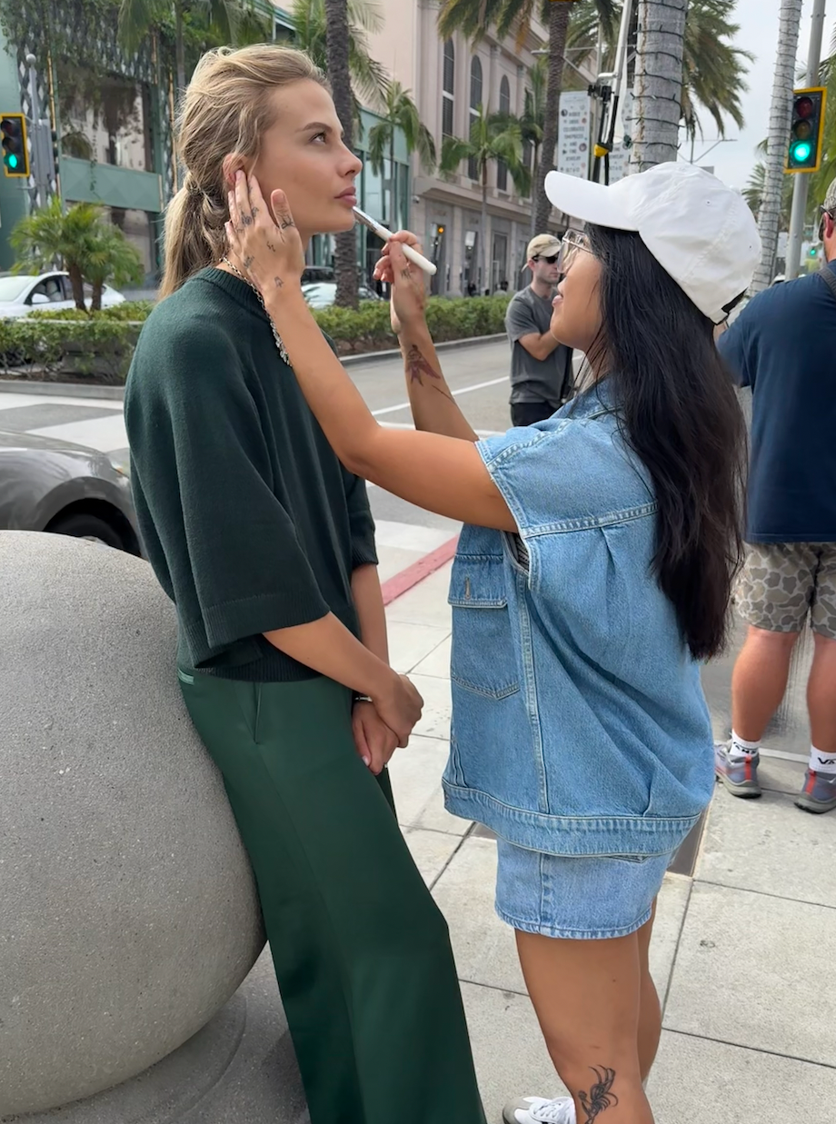 A woman with blonde hair and tattoos sits against a concrete structure while a makeup artist applies makeup to her face on a city street with palm trees and traffic lights in the background.