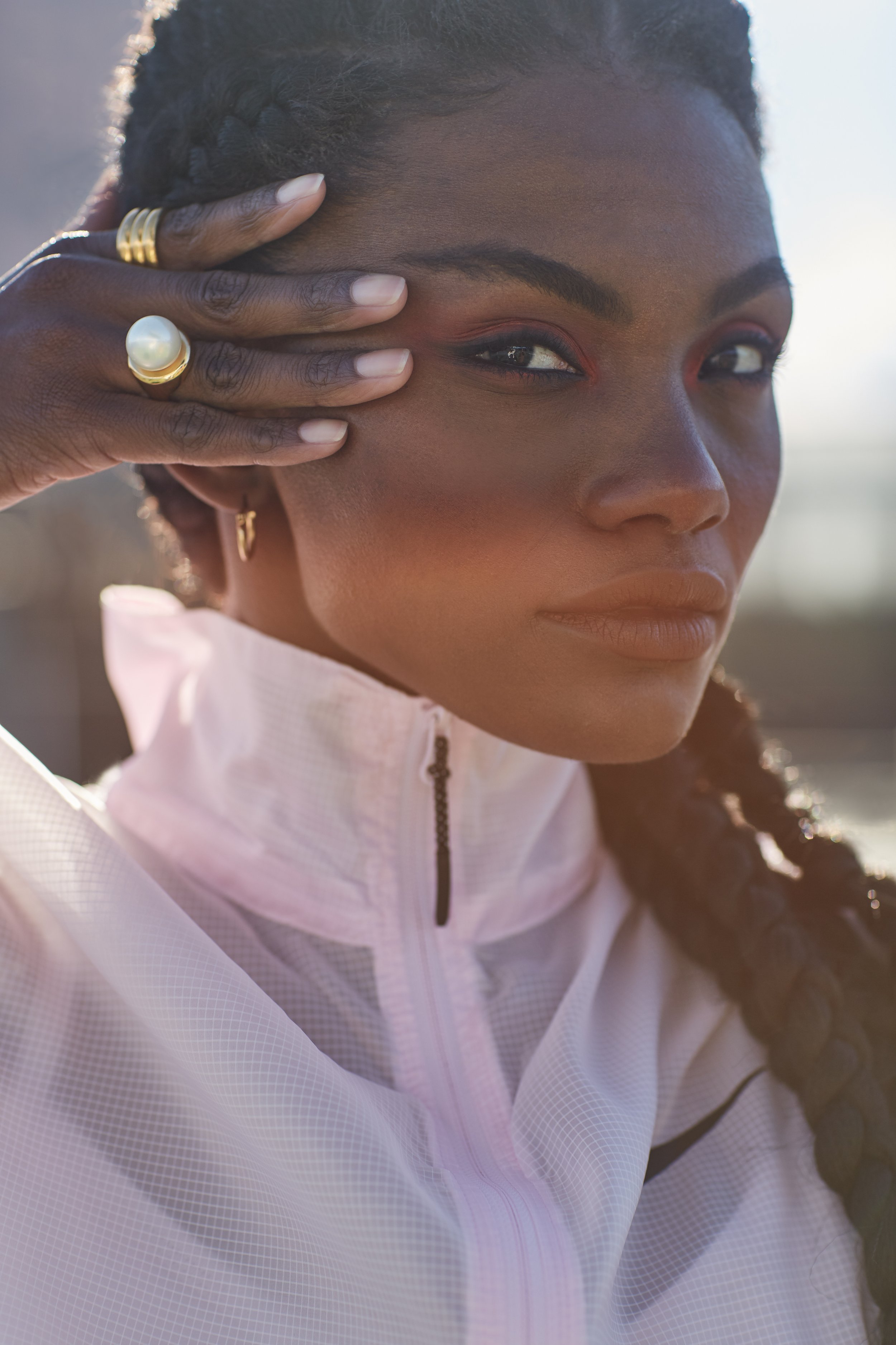 A woman with dark skin and braided hair holds up her hand with jewelry, wearing a white athletic jacket, outdoors with sunlight in the background.
