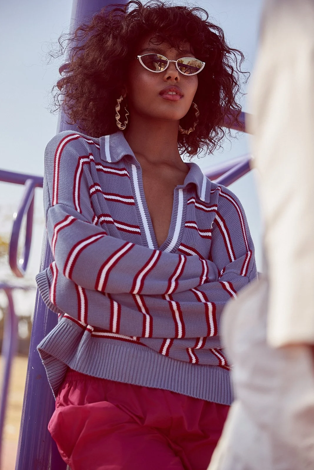 A woman with curly hair and sunglasses standing outdoors with a blue and white background, wearing a striped sweater and pink pants.