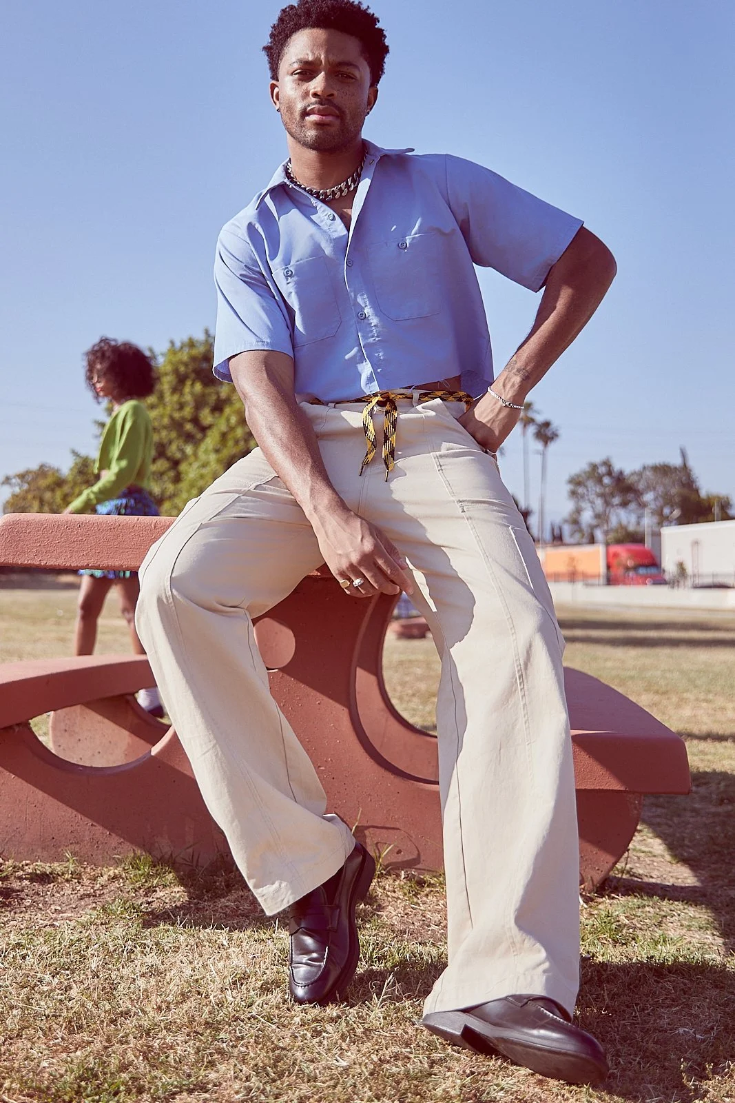 A young man with curly hair is sitting on a park bench, wearing a light blue short-sleeve shirt, cream-colored wide-legged pants, and black shoes. He is looking at the camera with a serious expression, with his right hand resting on his thigh and his