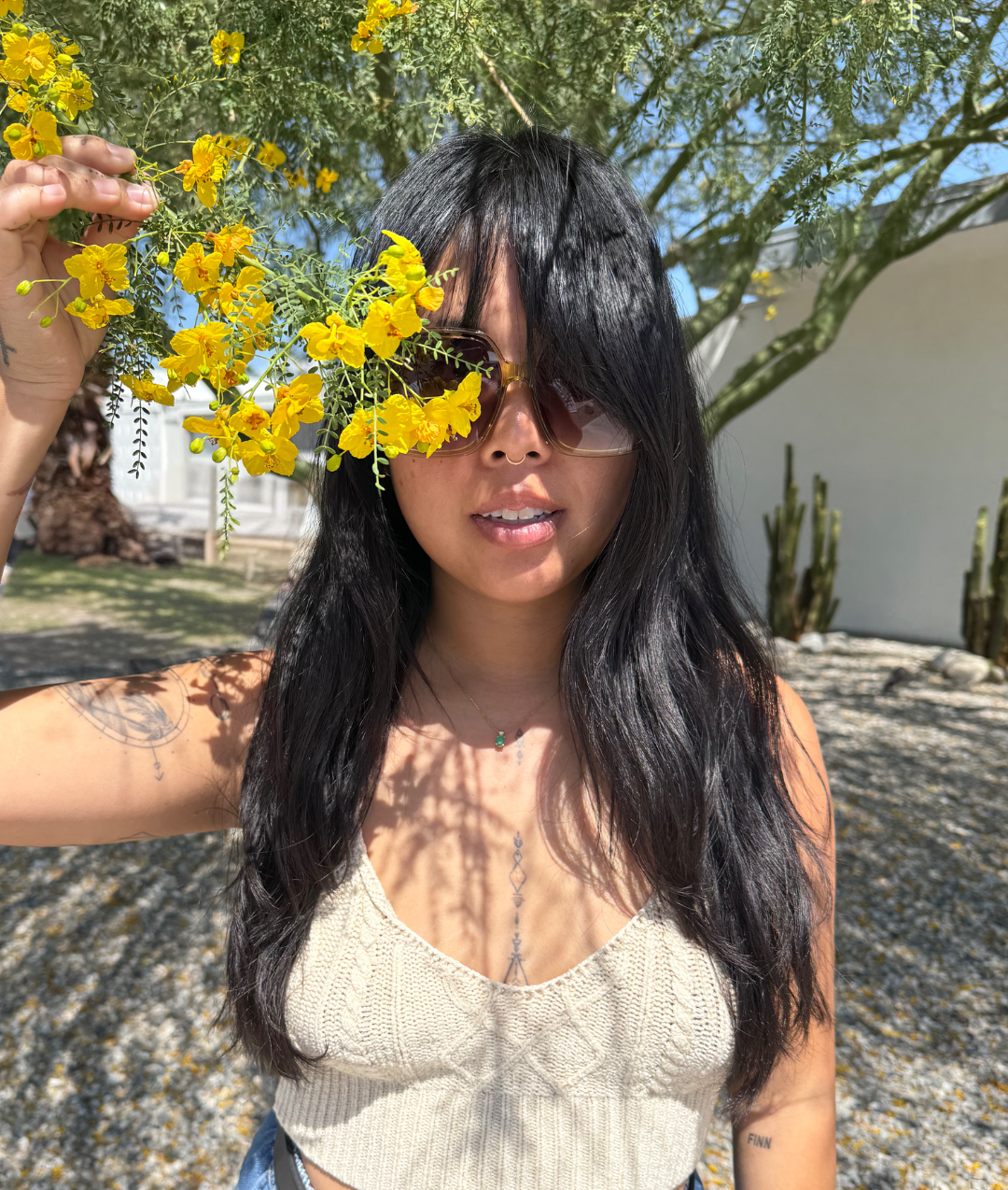 A woman with black hair wearing sunglasses and a beige sleeveless top standing outdoors under a tree with yellow flowers, holding a branch with yellow flowers next to her face.