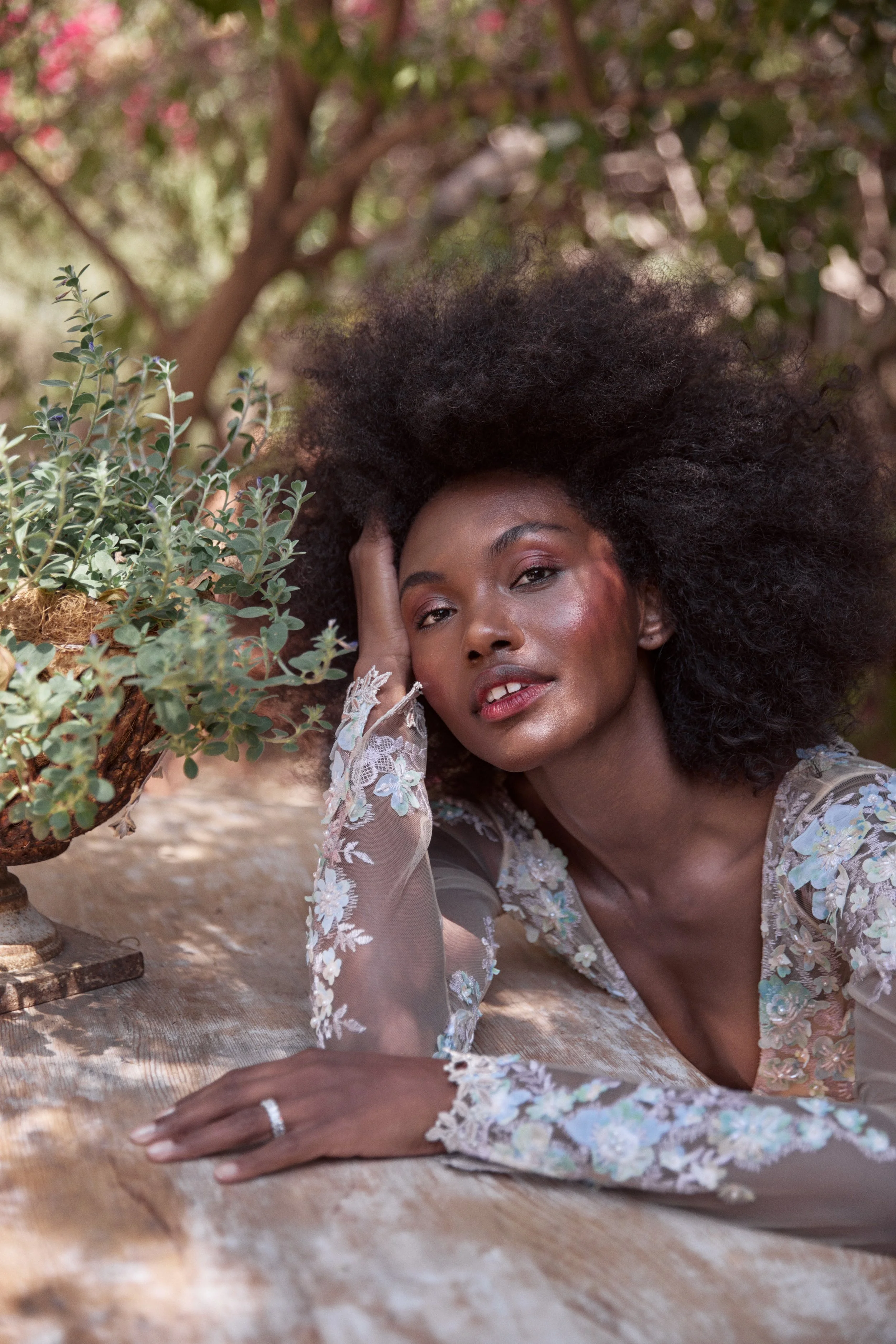 A woman with voluminous curly hair lying on a wooden table outdoors, resting her head on her hand, wearing a sheer, embroidered top with floral designs, surrounded by greenery and pink flowers.