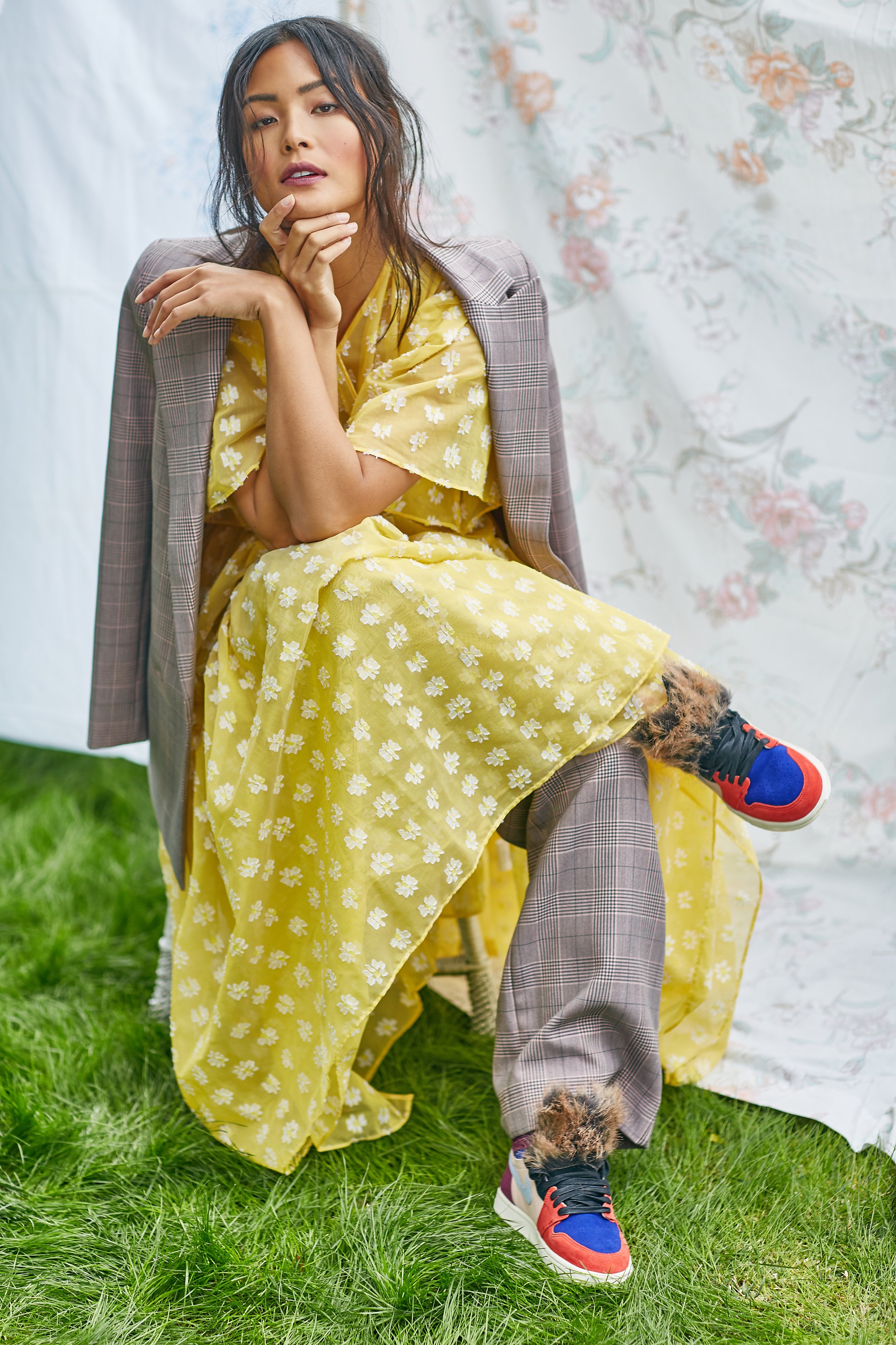 A woman dressed in a yellow floral dress and a plaid blazer sitting on a chair outdoors. She has dark, wavy hair and her hand resting near her chin. She is also wearing colorful sneakers with fur accents, and the background includes a floral curtain 