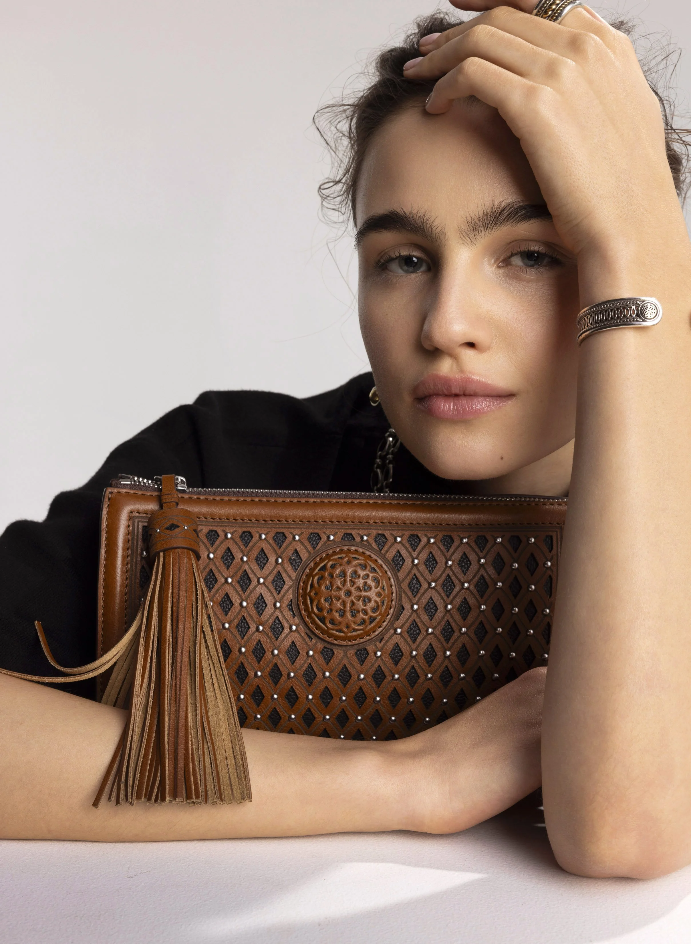 A woman with blonde hair, blue eyes, and light skin resting her head on her left hand, holding a brown patterned clutch with a tassel and silver studded accents, wearing a black top and silver jewelry.