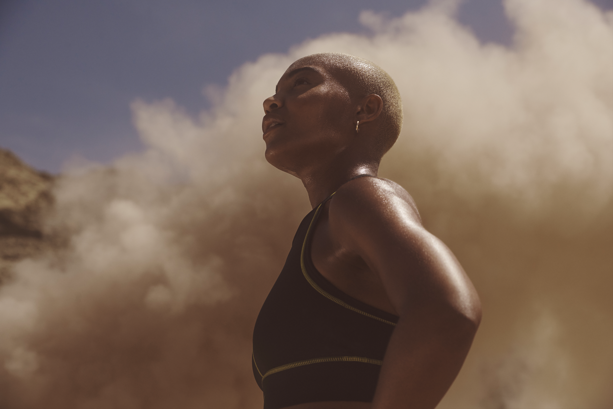 A woman with a shaved head and hoop earrings looks upward against a backdrop of sand and clouds.