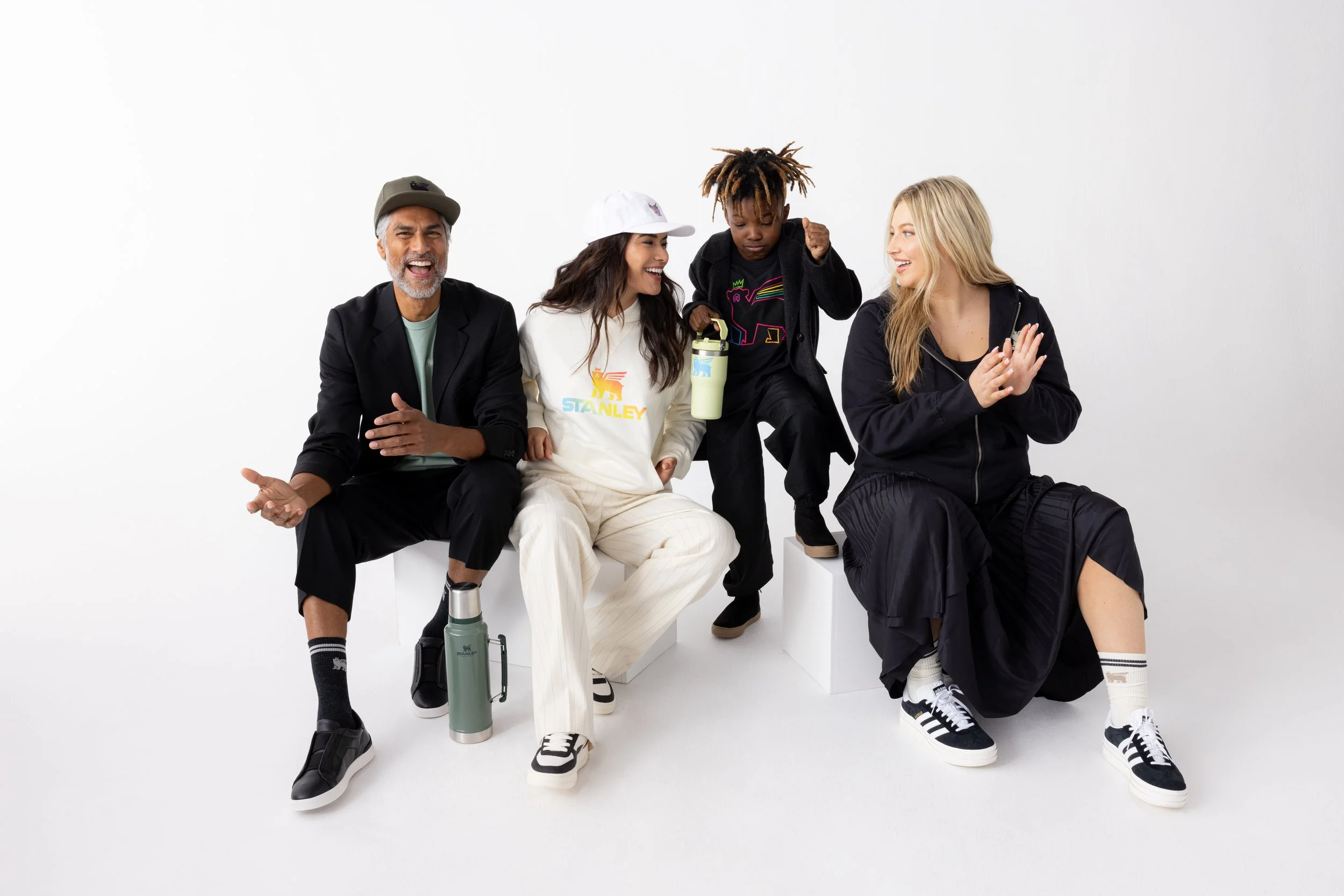 Four people sitting on white blocks against a plain white background, smiling and interacting. A man with gray hair and a beard, wearing a hat, black jacket, and pants, is laughing. Next to him, a woman in a white hoodie and pants, with long dark hai