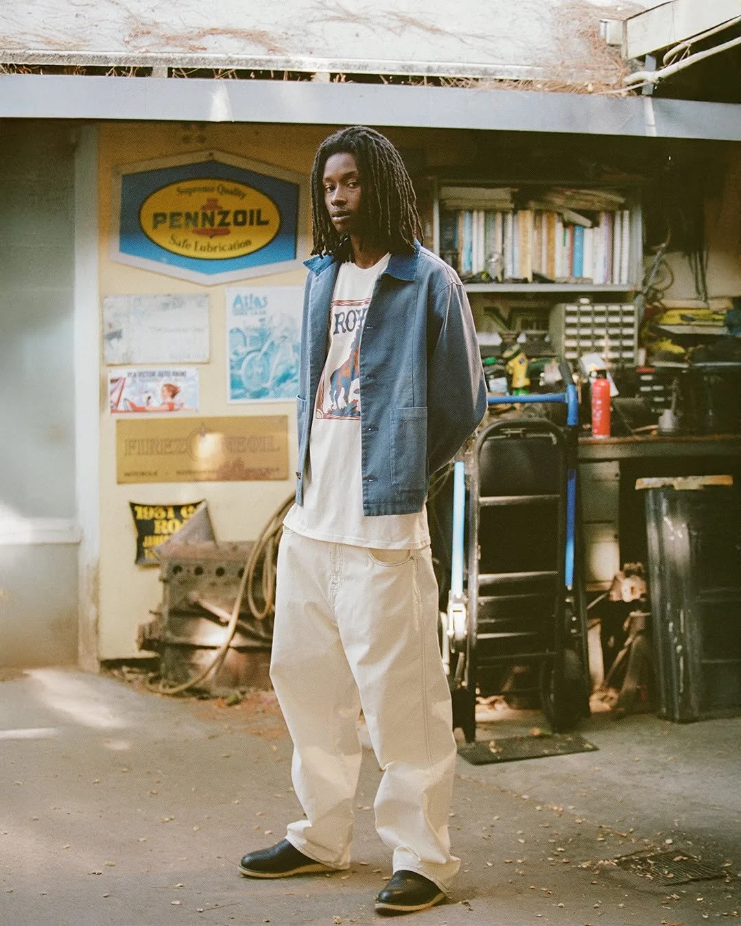 A young man with dreadlocks wearing a denim jacket, graphic T-shirt, and loose white pants standing in a garage or workshop area with tools and shelves in the background.