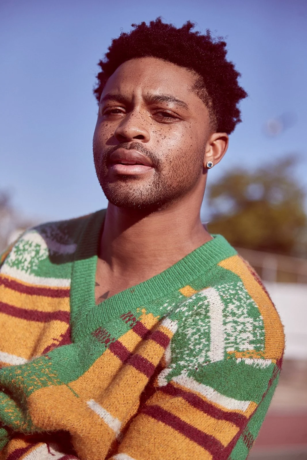 A young man with dark, curly hair and a beard, wearing a colorful, patterned sweater, standing outdoors in bright sunlight with a blurred background of trees and sky.