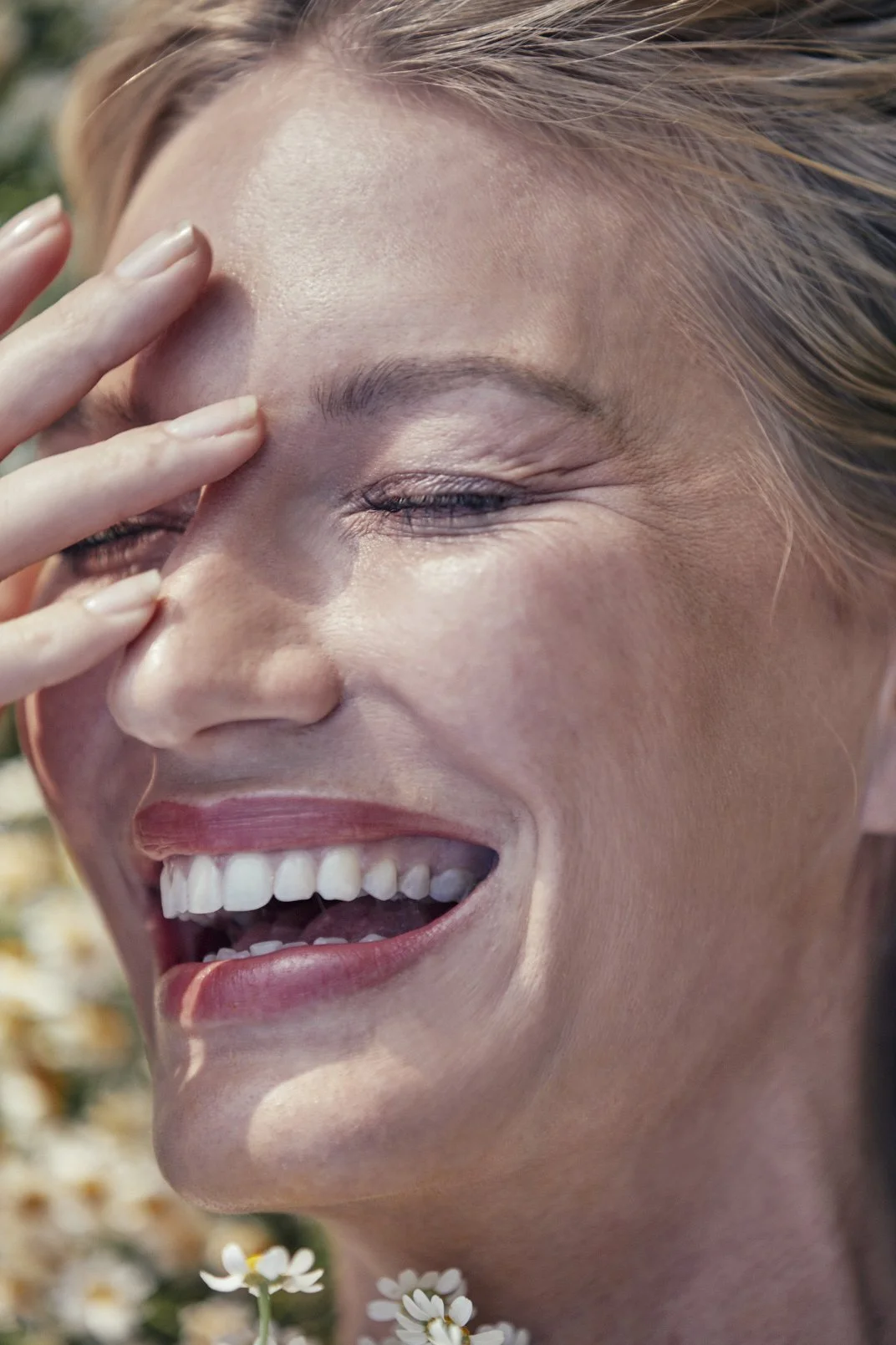Close-up of a smiling woman with her eyes closed, her hand touching her face, and surrounded by small white flowers.