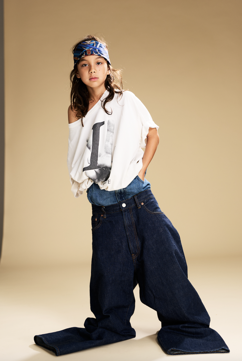 A young girl with long dark hair wearing a blue bandana, a loose white t-shirt with a large black 'L' printed on it, and oversized dark blue jeans against a plain beige background.