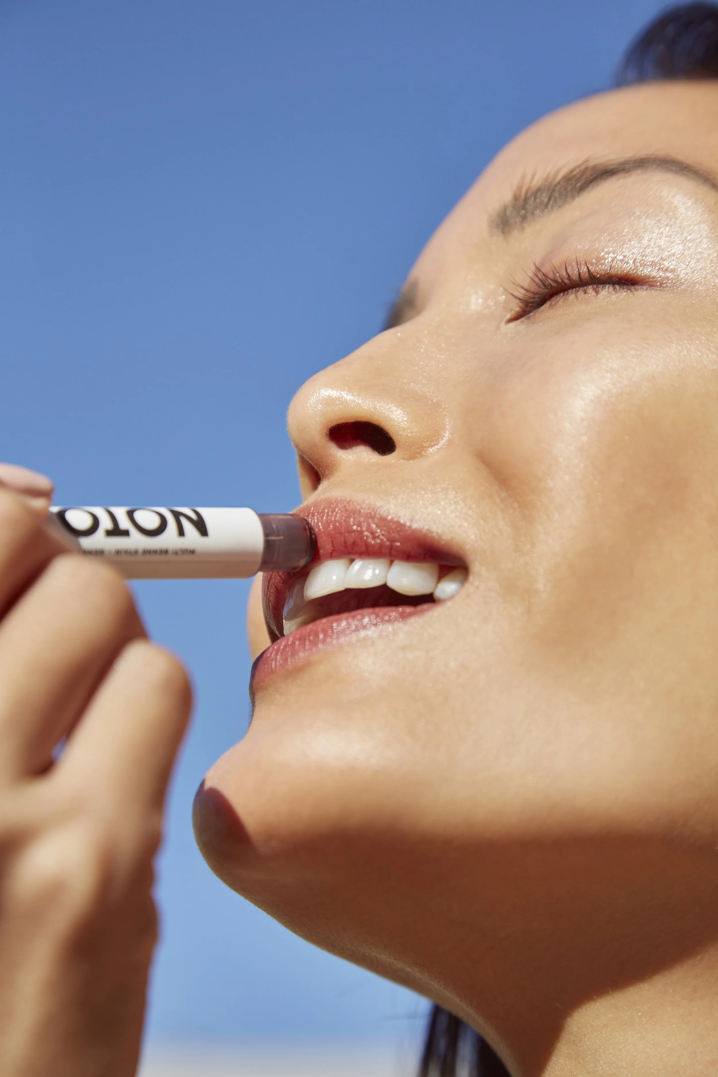 A woman with smooth skin and closed eyes applies lip balm outdoors against a blue sky.