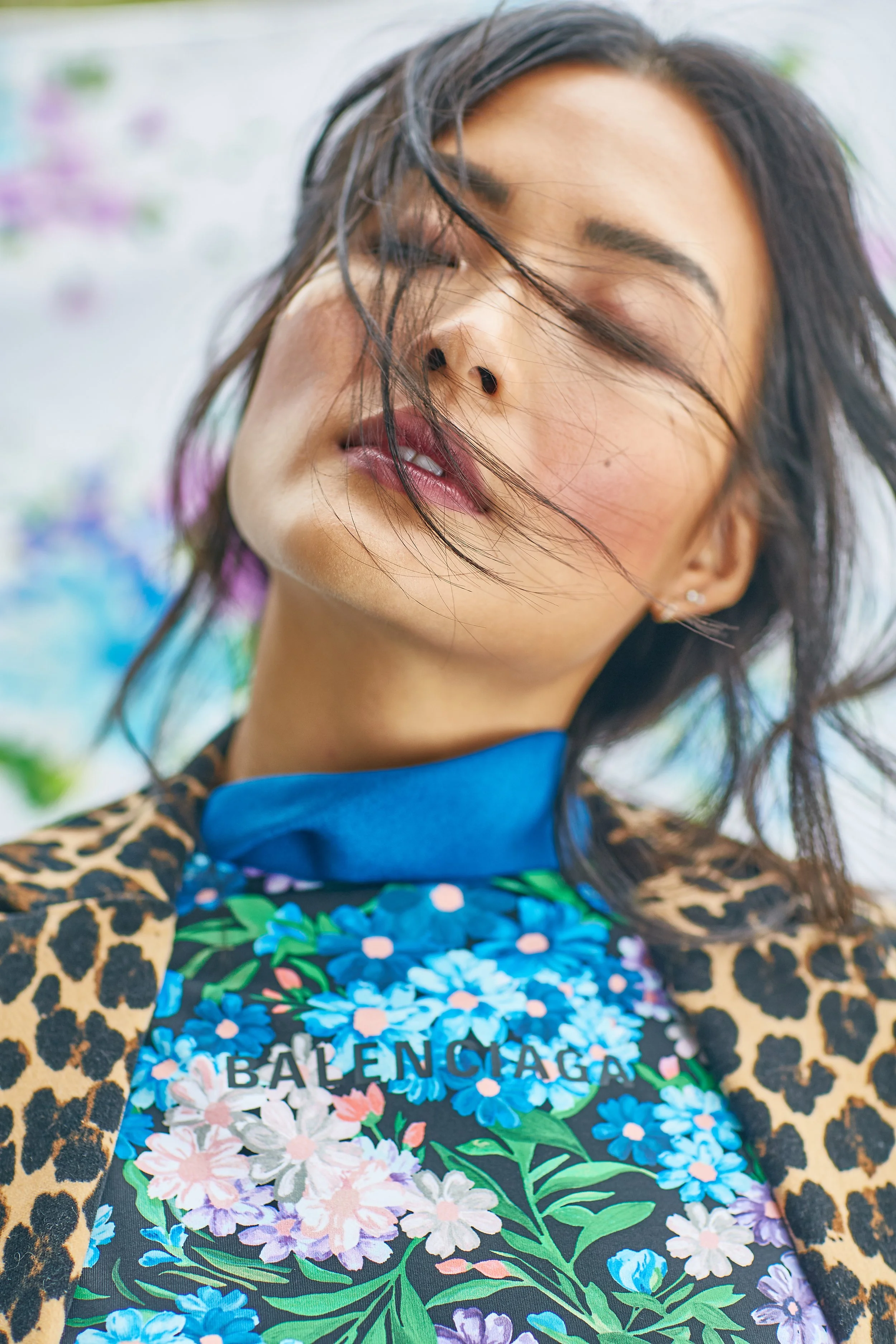 Close-up of a woman with messy hair, closed eyes, and partially open lips, wearing a colorful floral top and a leopard print jacket, with a blurred colorful background.
