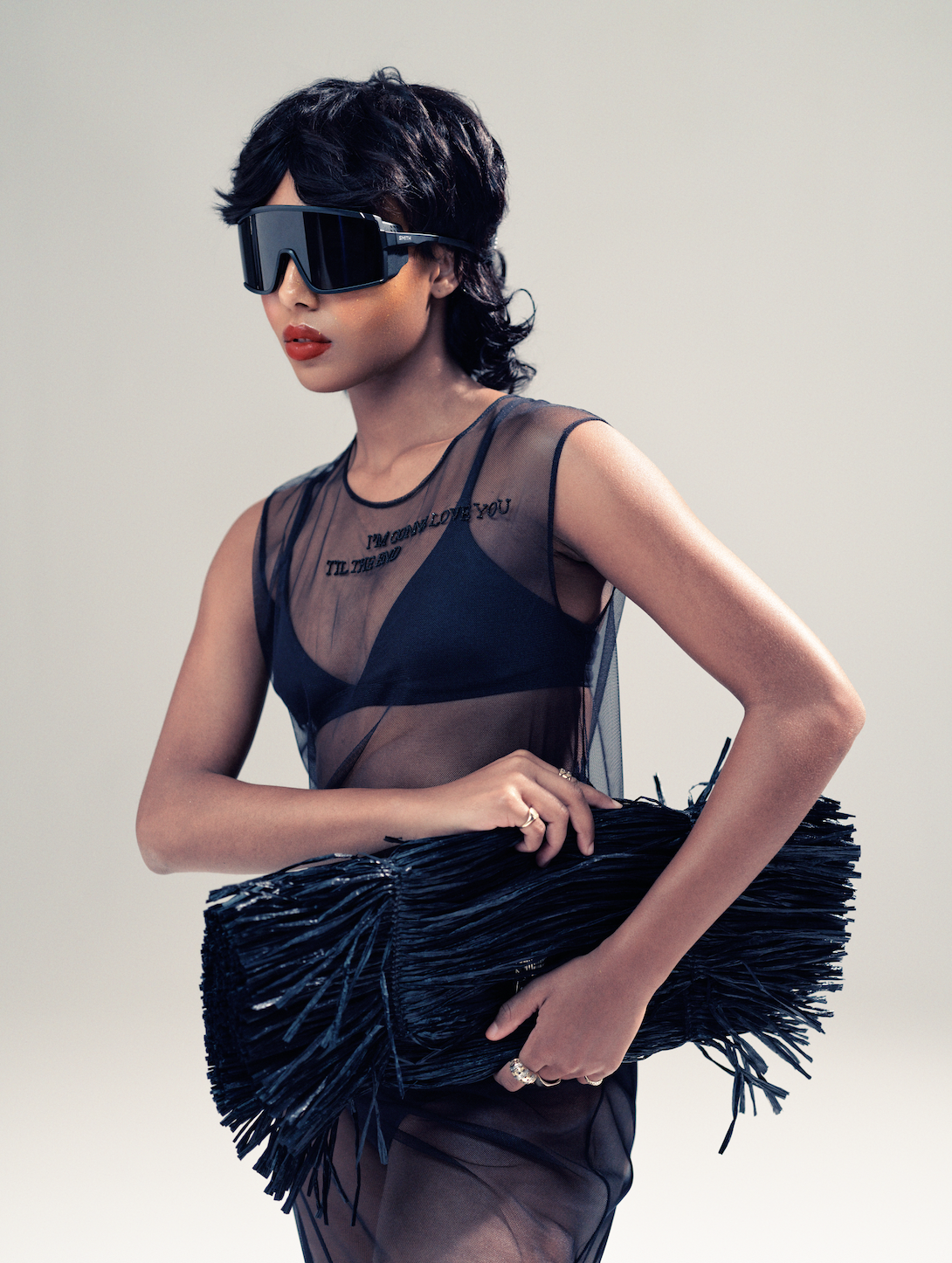A woman with short dark hair wearing large black sunglasses, a sheer sleeveless black top with embroidered text, holding a textured black handbag against a plain background.