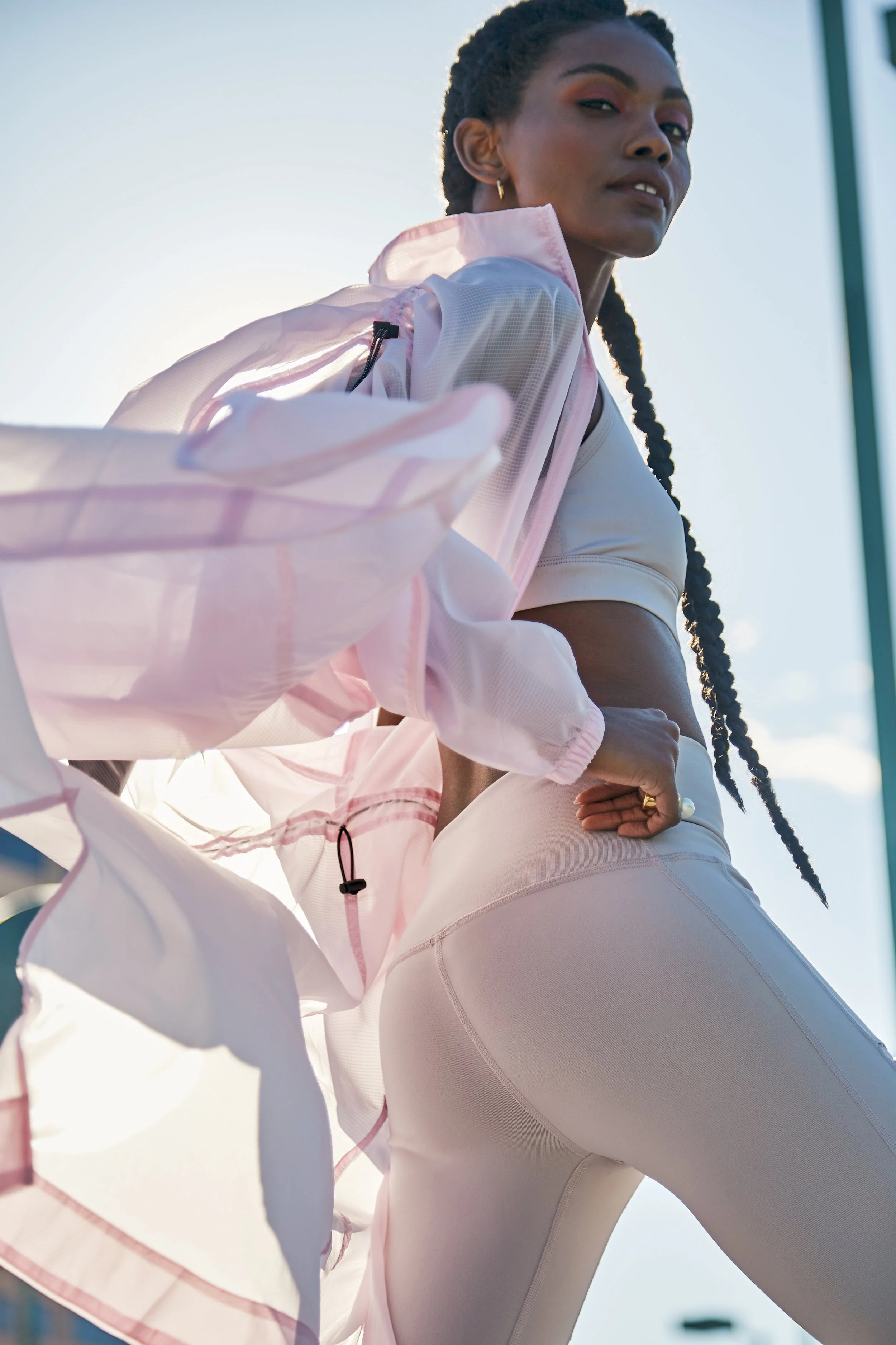 A young woman with long braided hair, wearing a light-colored sports bra, form-fitting athletic leggings, and a pink windbreaker, standing outdoors with a bright sky and sunlight in the background.
