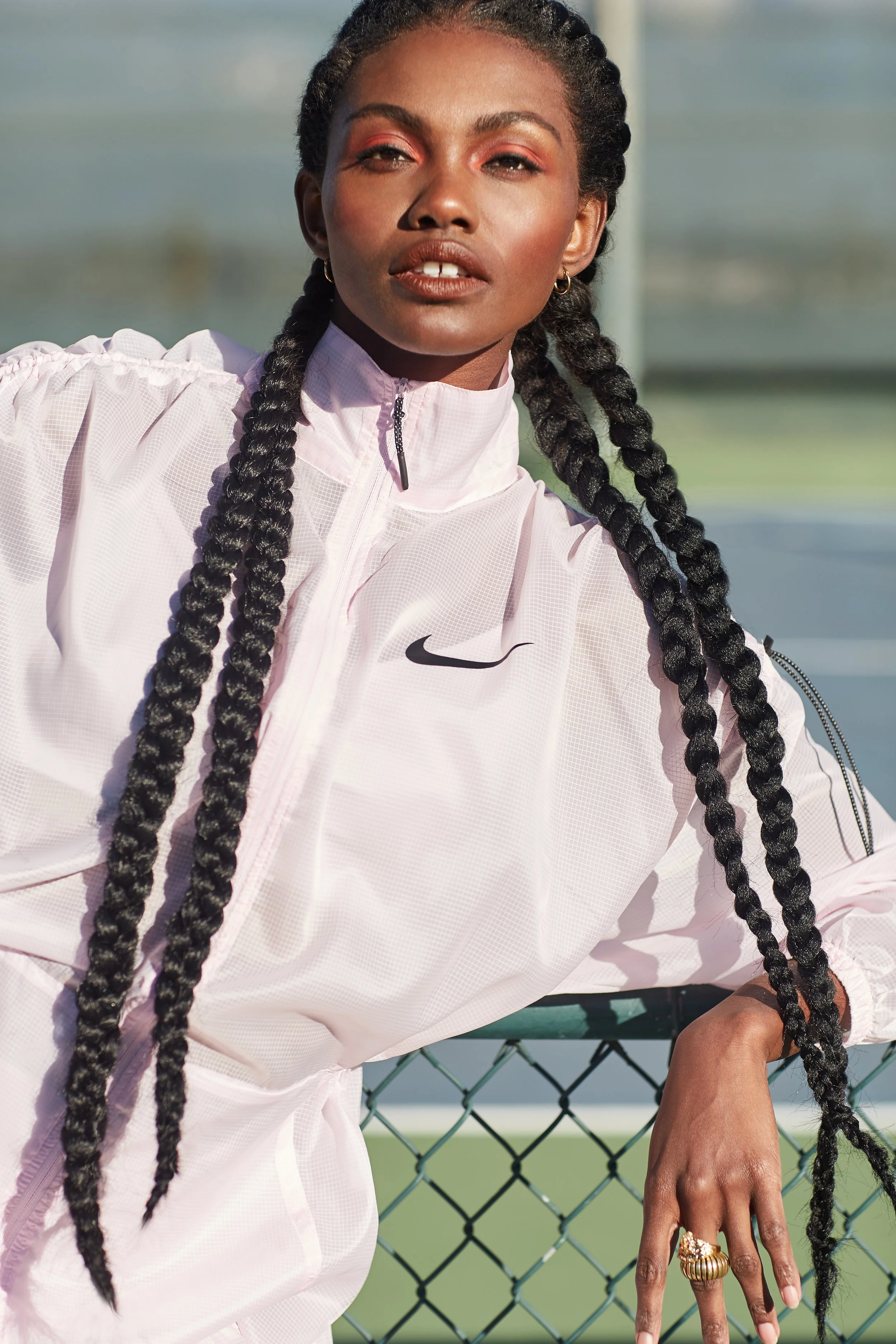 A woman with long braided hair wearing a light pink Nike jacket, standing outdoors by a chain-link fence.