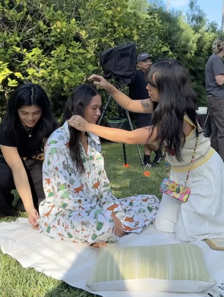 Young woman sits cross-legged on a blanket outdoors with two others nearby, having her makeup done by another woman, holding a brush in front of her. There is a camera crew in the background.