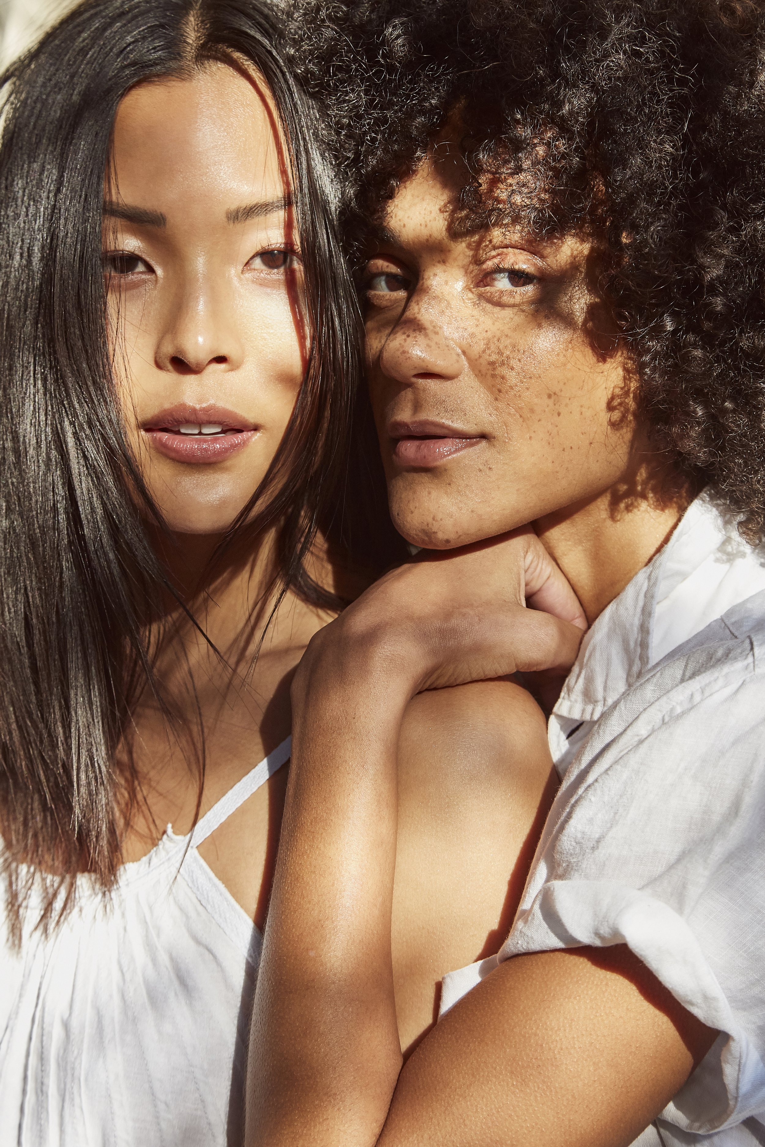 Close-up of two women with diverse skin tones and hair types, posing together outdoors in sunlight, wearing white tops.