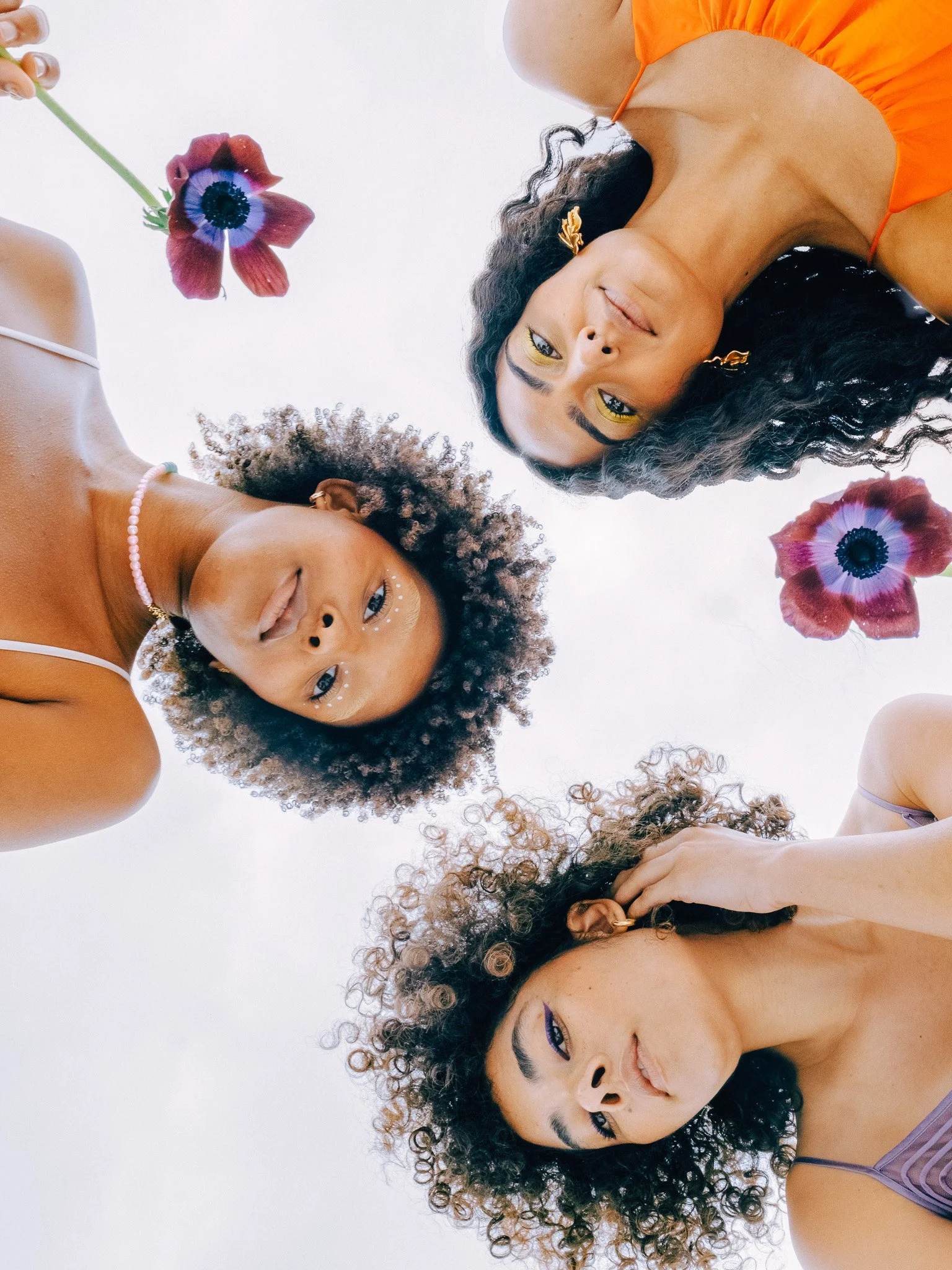 Three women with curly hair and unique makeup, looking down at the camera, with two purple and red flowers floating in the background.