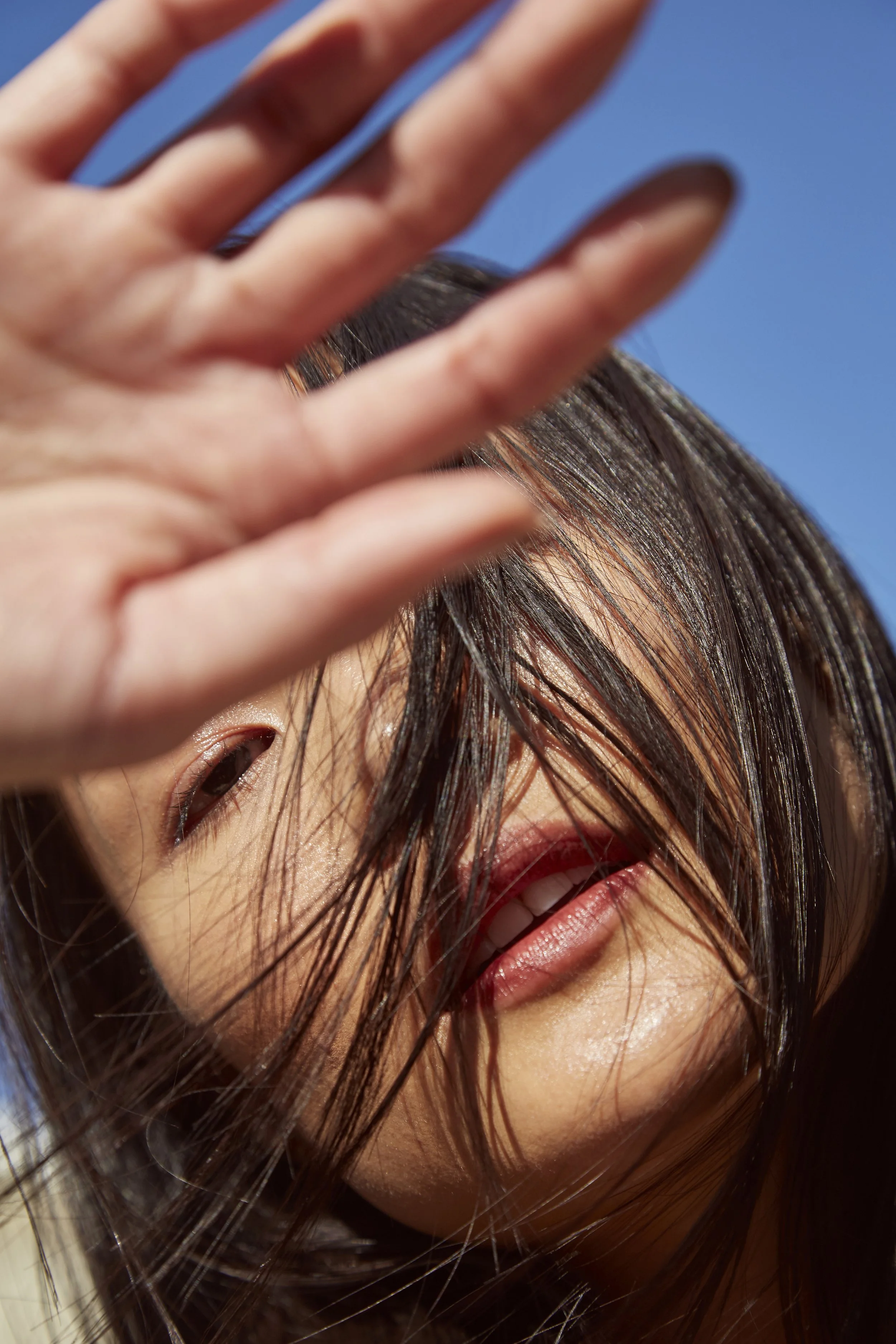 A smiling woman outdoors blocking part of her face with her hand, with long dark hair in front of her face, against a clear blue sky.