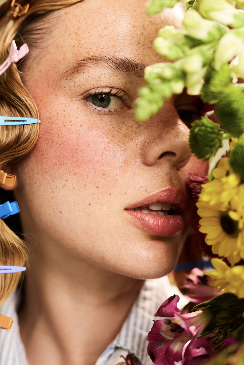 Close-up of a young woman's face with makeup and freckles, partially obscured by colorful flowers on one side.
