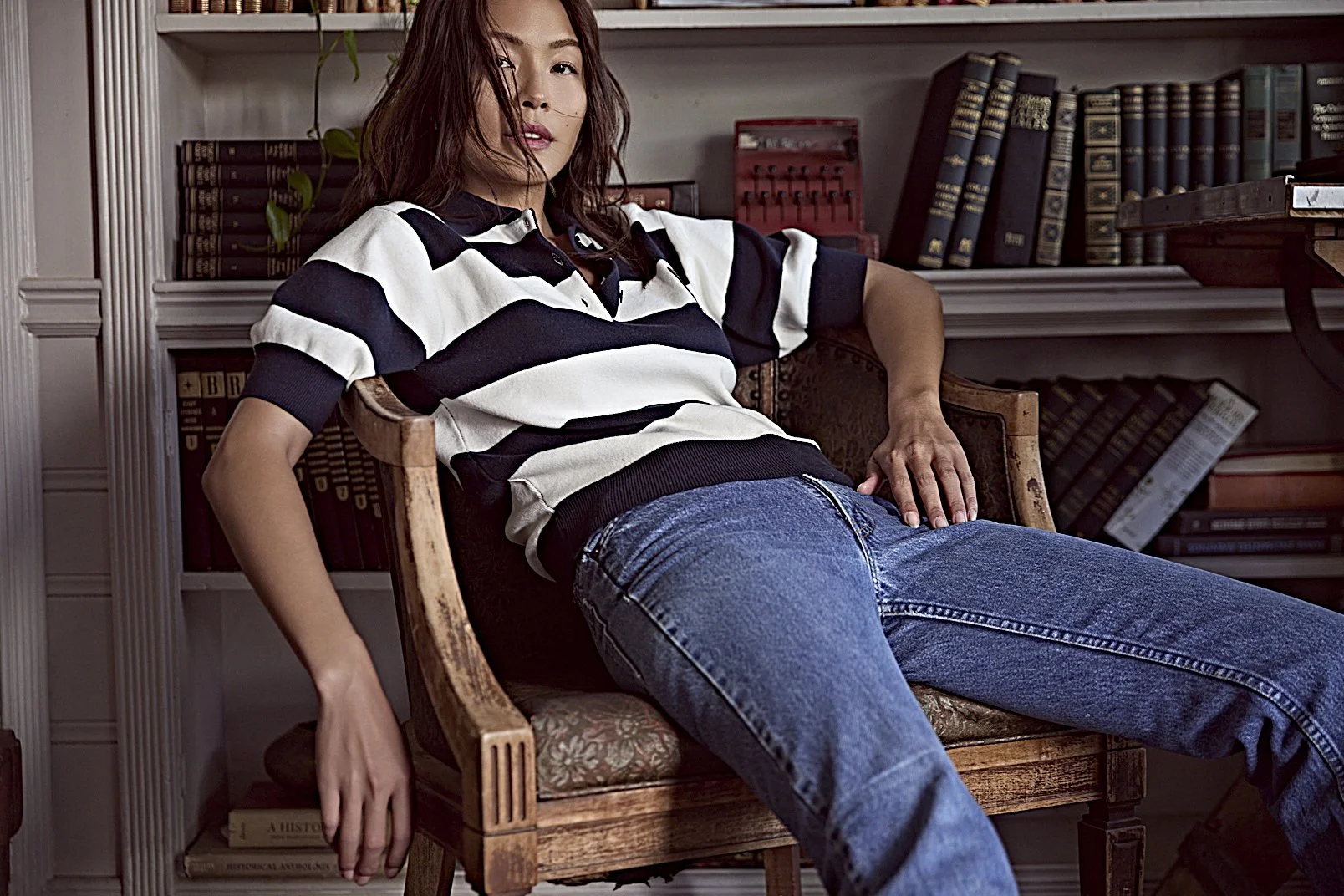 A woman reclining on a vintage wooden chair in a room filled with books, wearing a striped polo shirt and jeans.