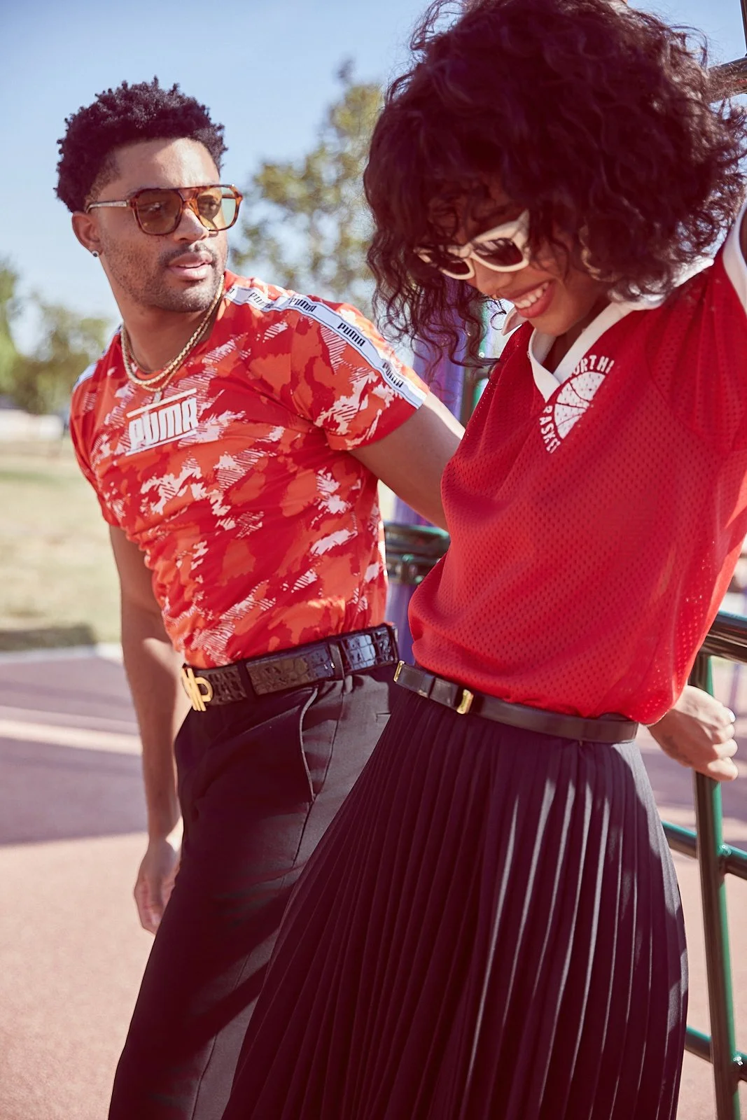 A man and a woman are outdoors, both wearing sunglasses and red sports apparel. The man has short curly hair, a beard, and is wearing a patterned red Puma shirt with graphics, black pants, and a belt. The woman has voluminous curly hair, is smiling, 